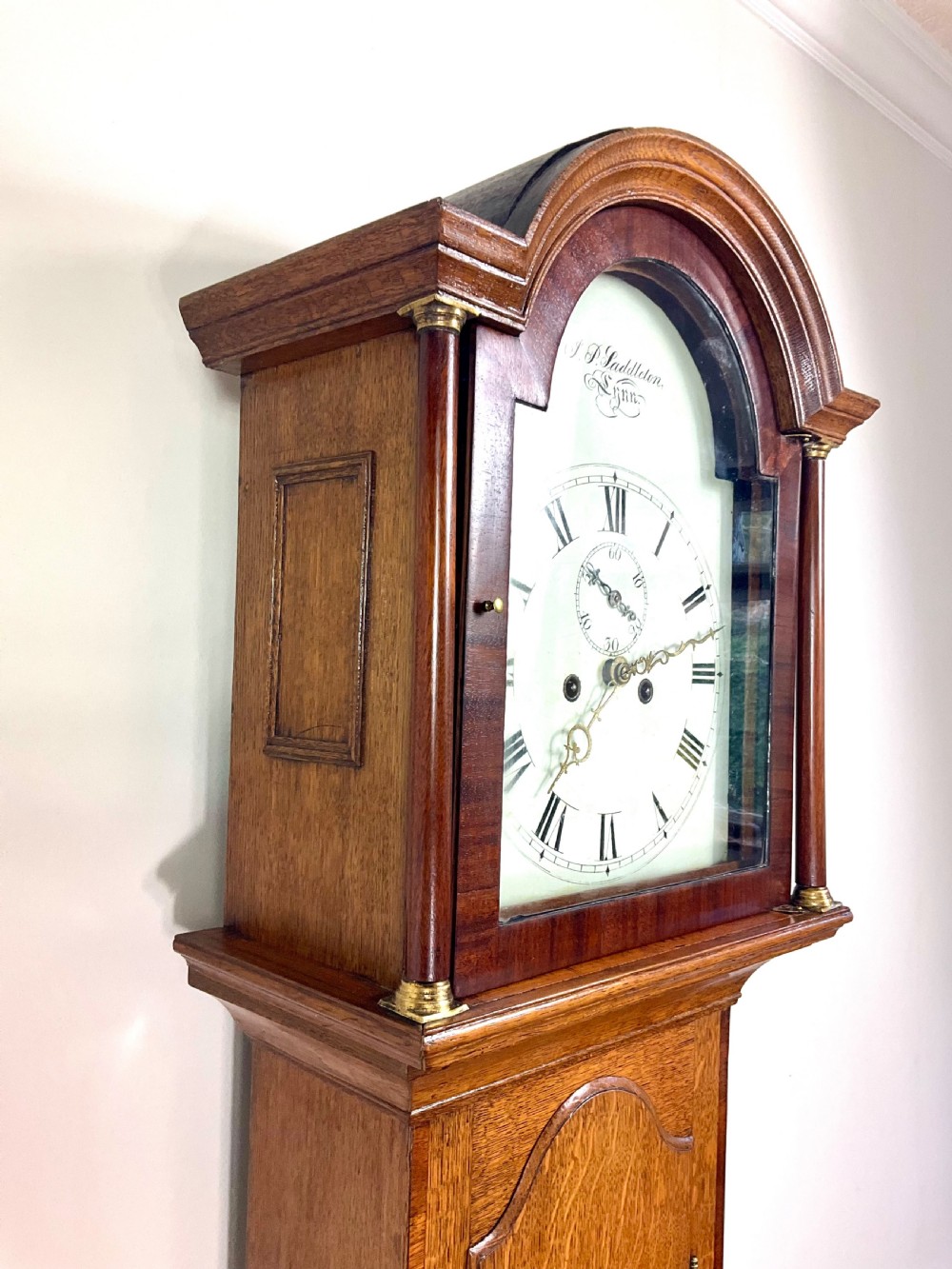 An antique Georgian longcase clock hood crafted from light oak with an arched glass door, brass-capped side columns, and a painted white dial with Roman numerals.