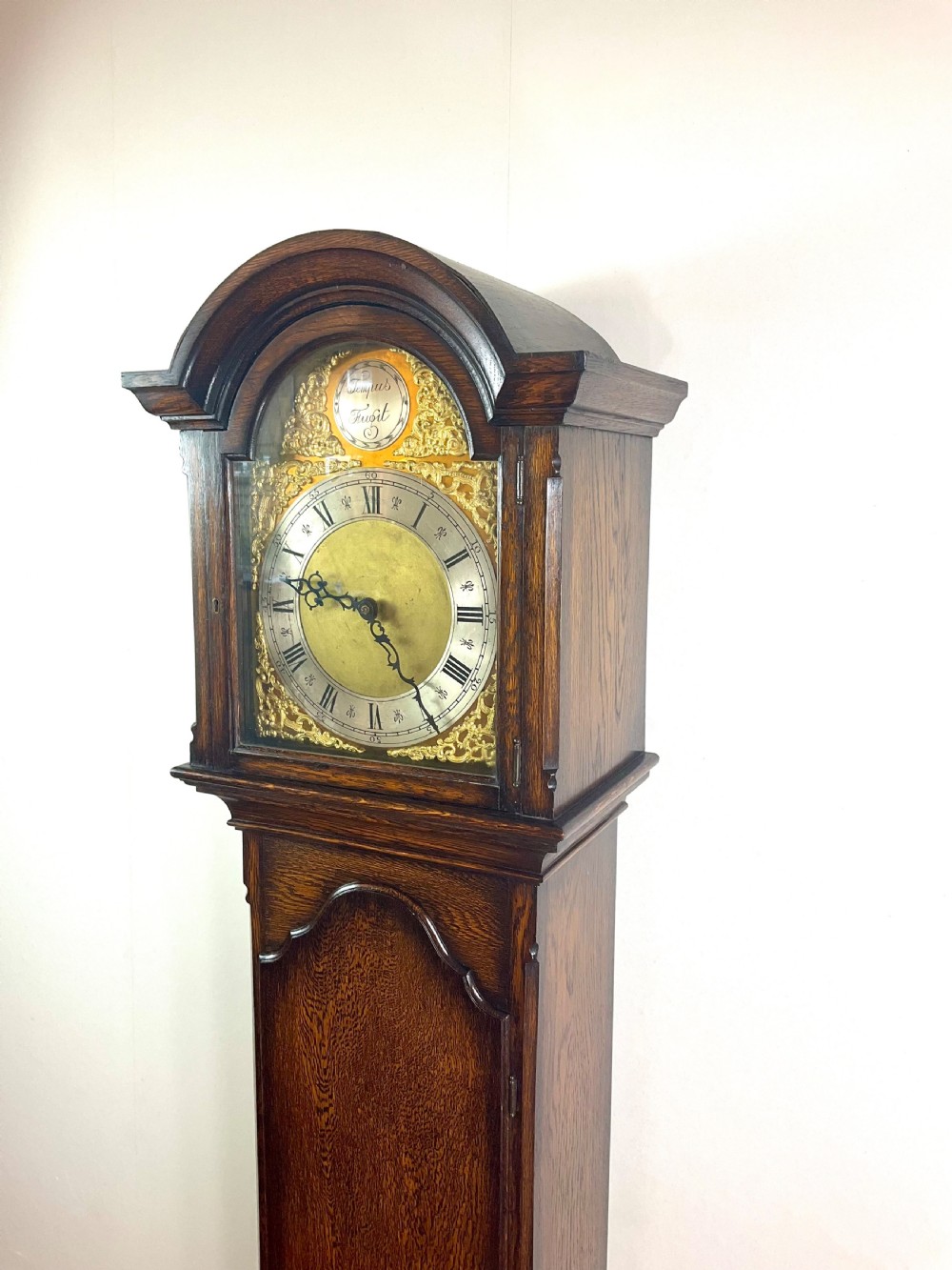 A dark oak grandmother longcase clock featuring an arched hood, clear glass door, and a brass dial with Roman numerals.