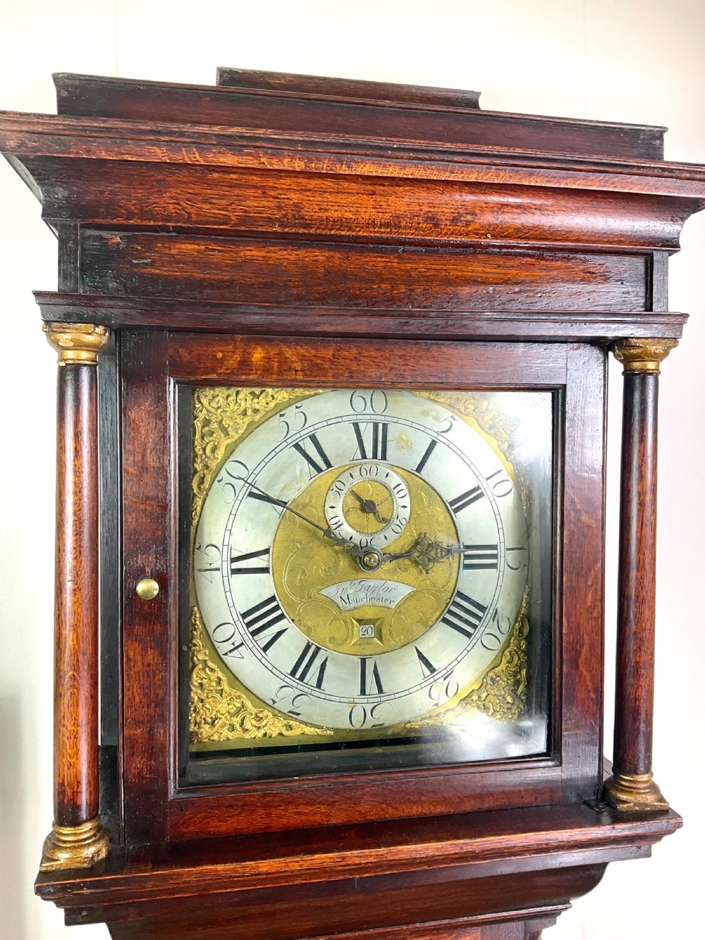 An Early Georgian longcase clock constructed from dark oak featuring a square hood, brass dial with Roman numerals, and an arched trunk door. View IV