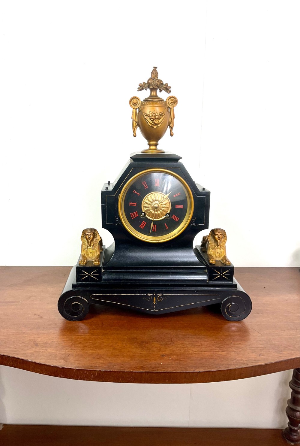 A black slate Egyptian Revival mantel clock featuring a central dial with red Roman numerals, flanked by two gilt bronze sphinxes and topped with a gilt urn finial.