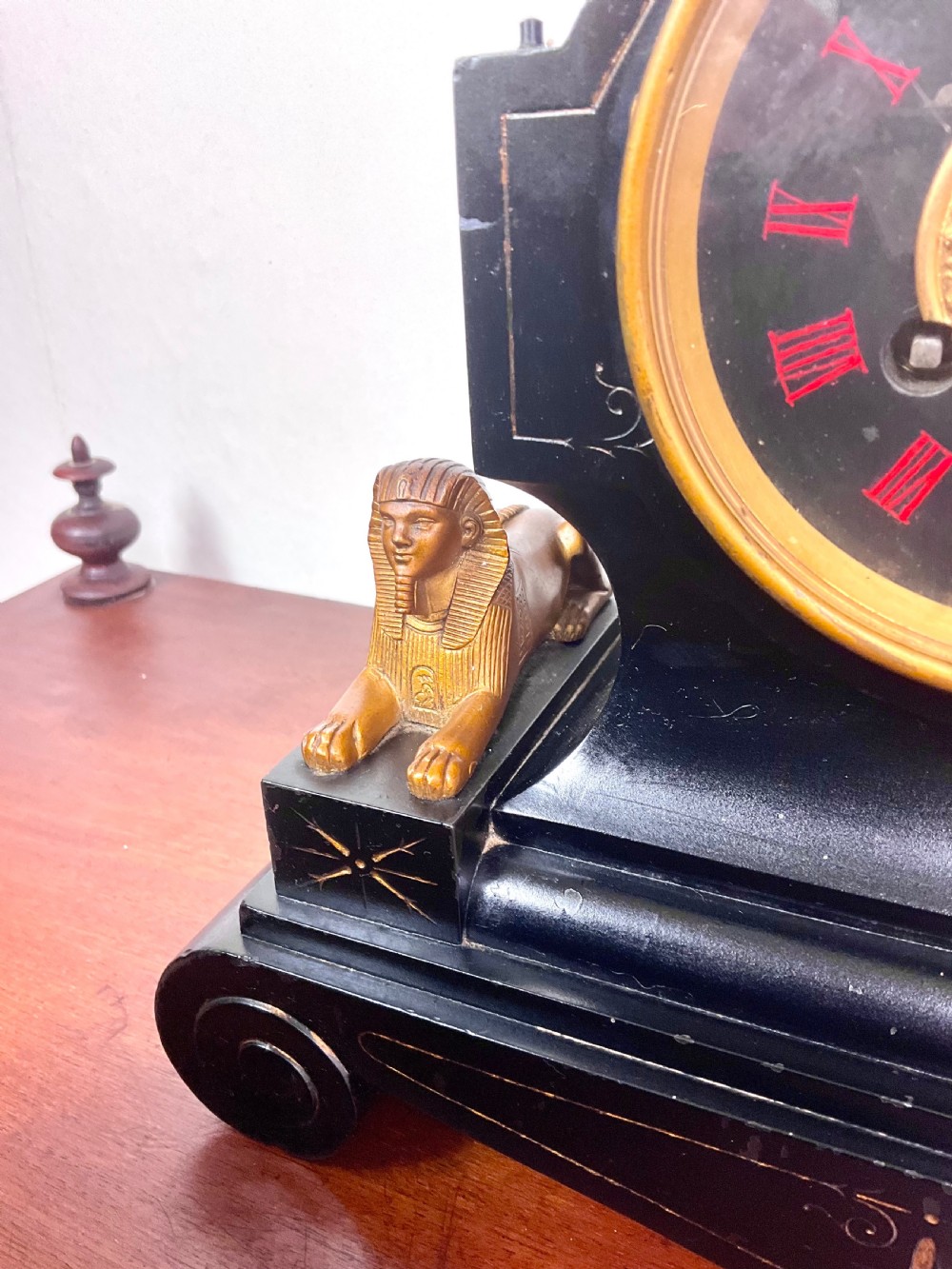 A black slate Egyptian Revival mantel clock featuring a central dial with red Roman numerals, flanked by two gilt bronze sphinxes and topped with a gilt urn finial. View V
