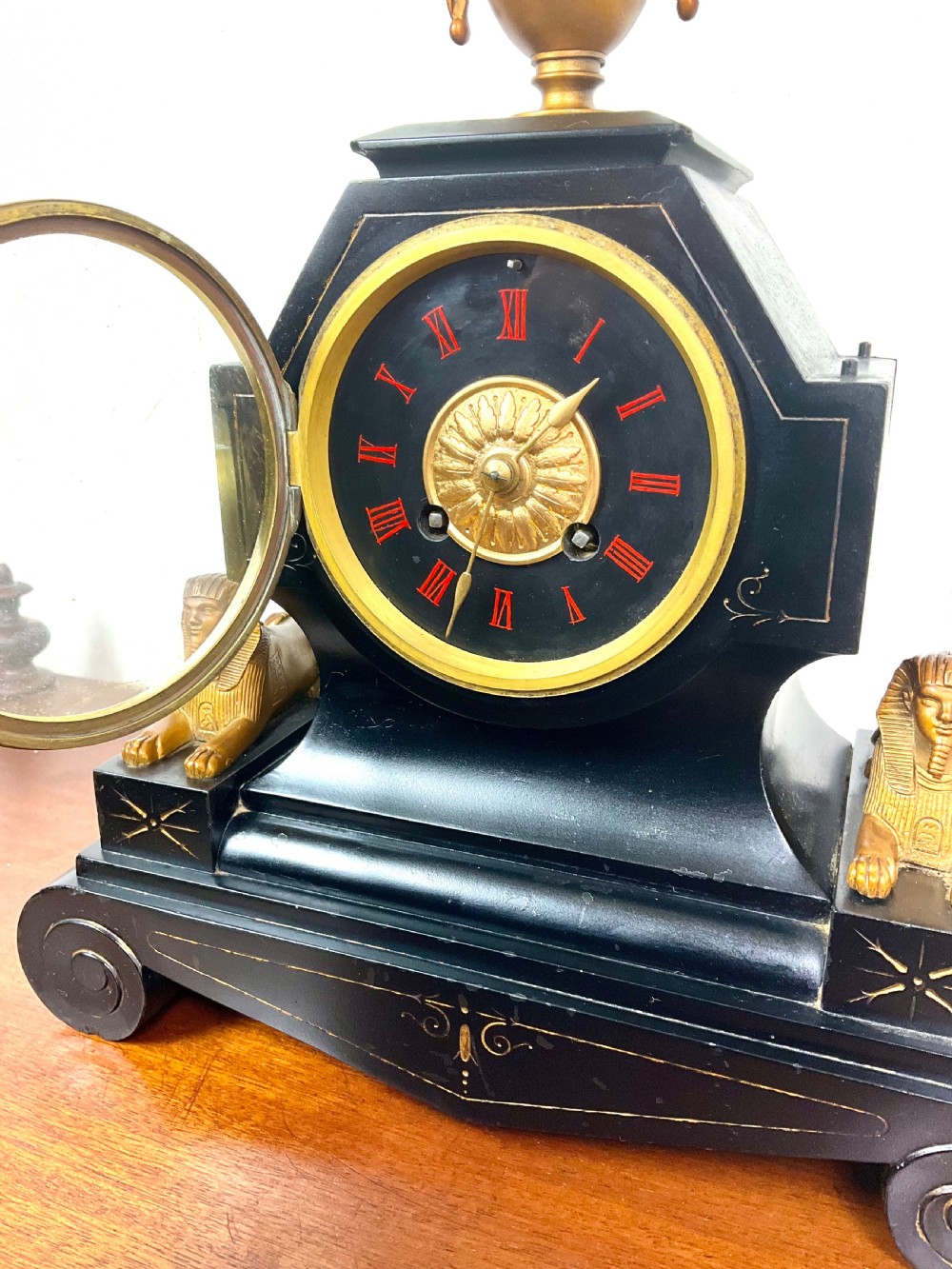 A black slate Egyptian Revival mantel clock featuring a central dial with red Roman numerals, flanked by two gilt bronze sphinxes and topped with a gilt urn finial. View VIII
