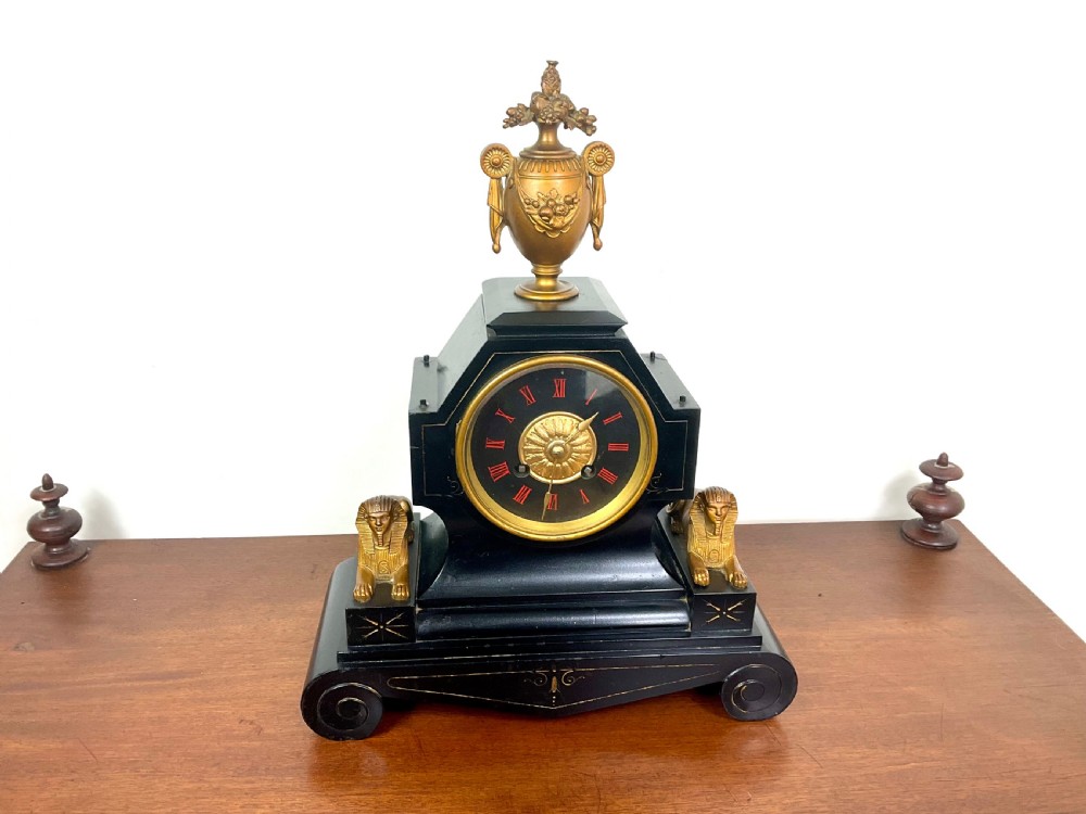 A black slate Egyptian Revival mantel clock featuring a central dial with red Roman numerals, flanked by two gilt bronze sphinxes and topped with a gilt urn finial. View X