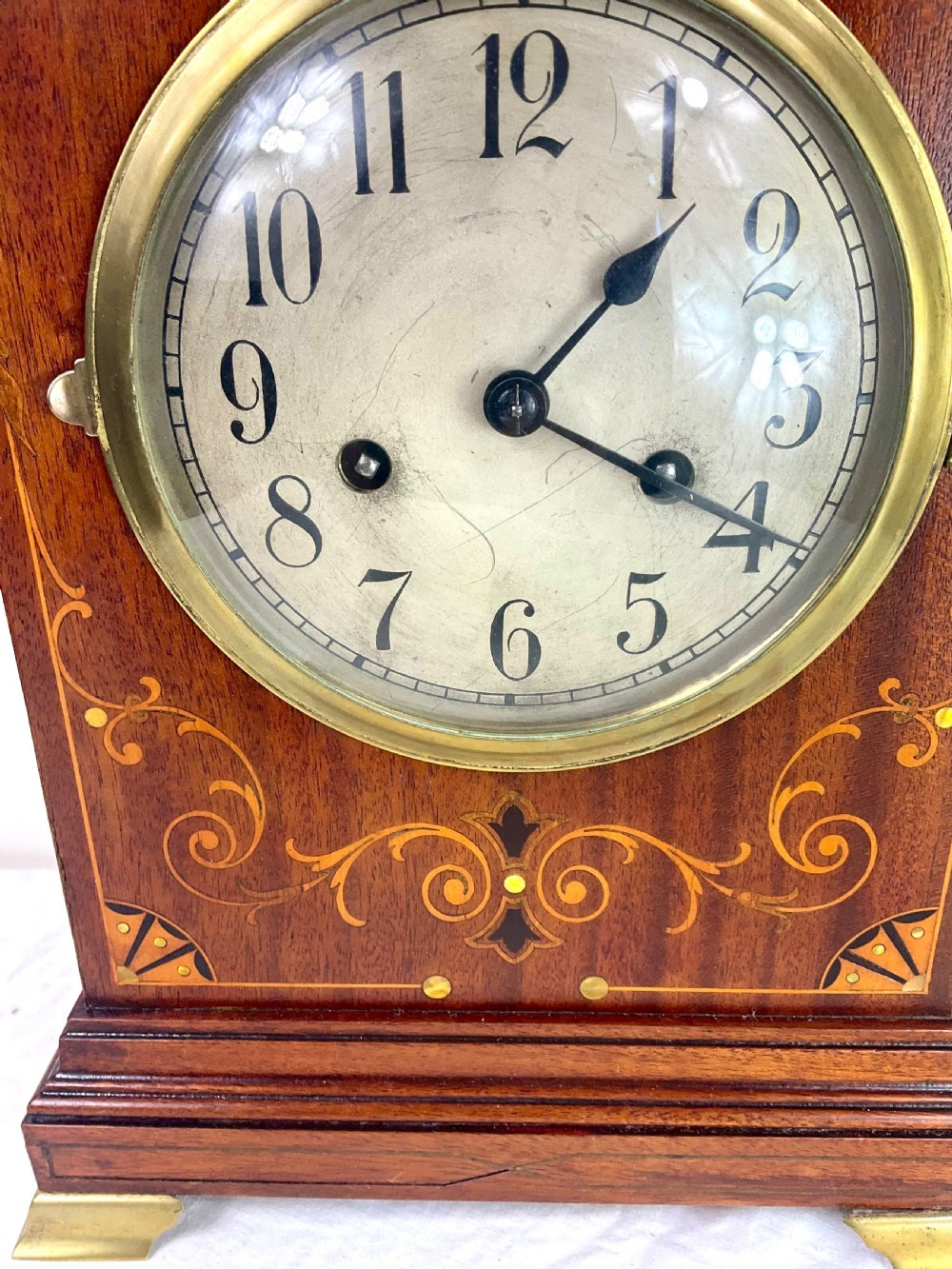 An Edwardian inlaid mahogany mantel clock featuring a circular dial, brass bezel, floral marquetry detailing, and brass bracket feet. View II