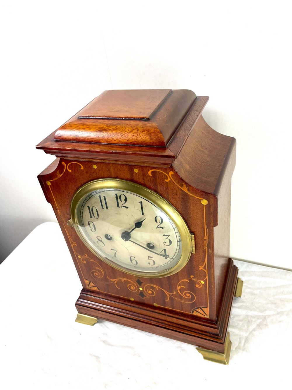 An Edwardian inlaid mahogany mantel clock featuring a circular dial, brass bezel, floral marquetry detailing, and brass bracket feet. View VI