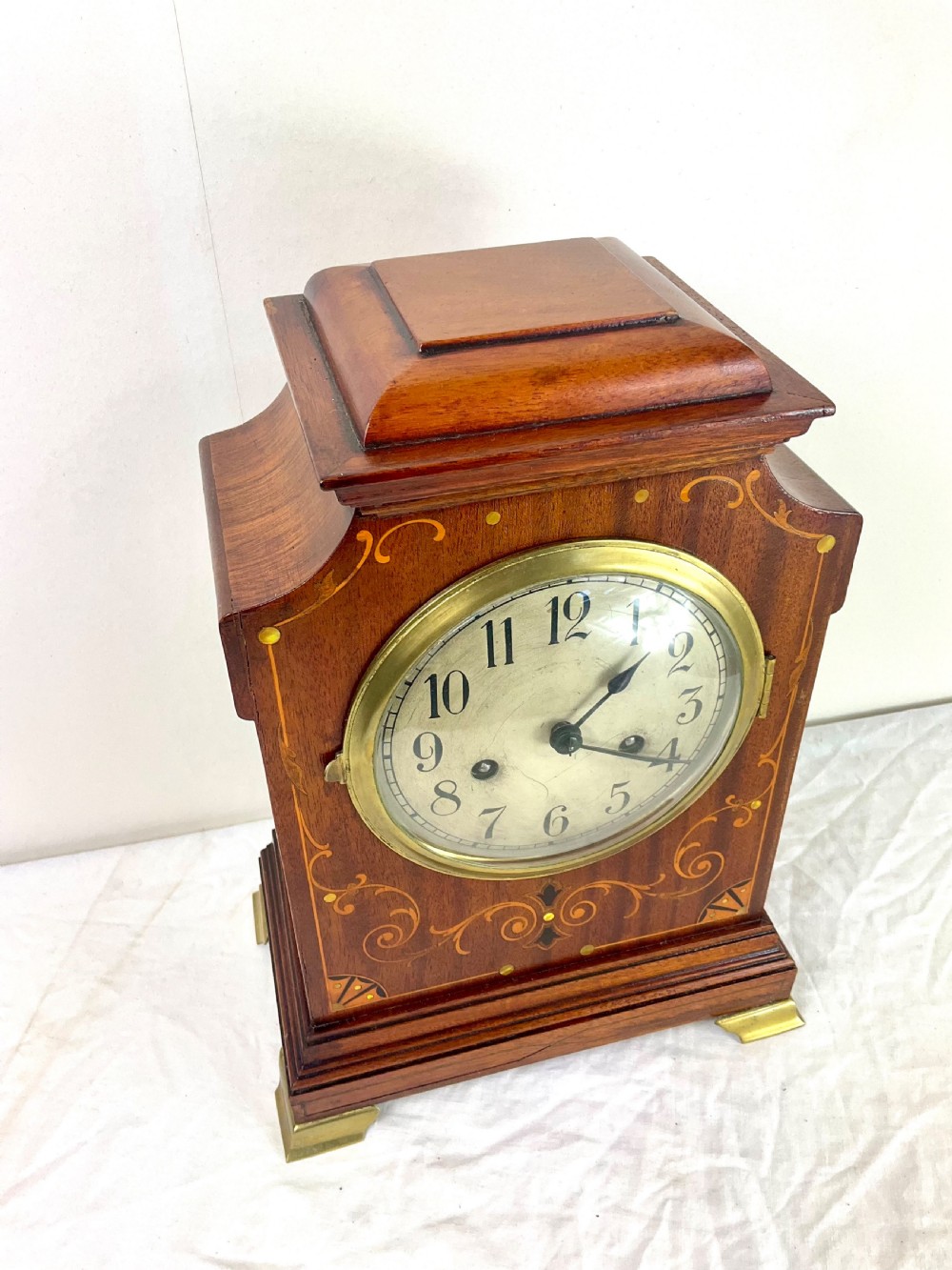An Edwardian inlaid mahogany mantel clock featuring a circular dial, brass bezel, floral marquetry detailing, and brass bracket feet. View VII