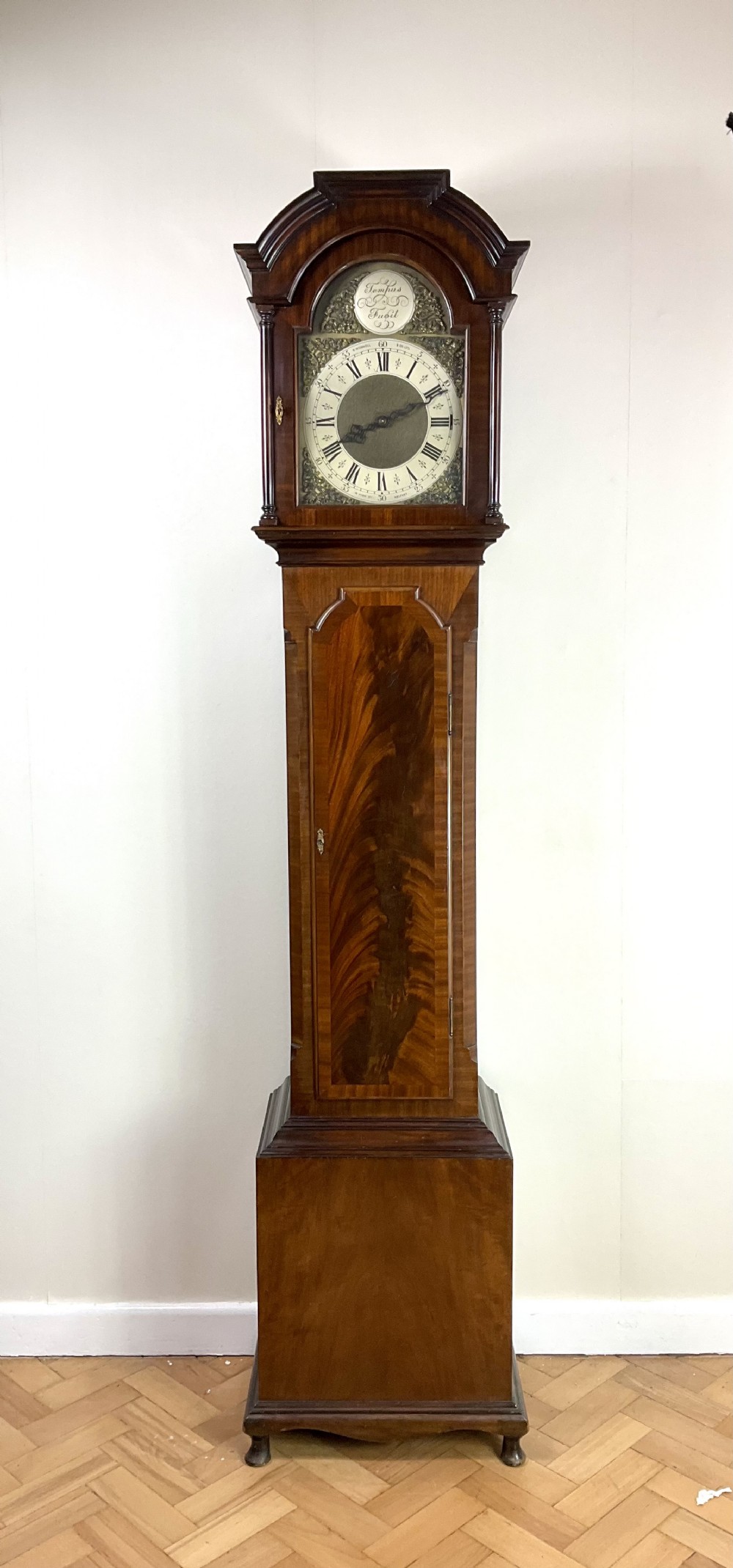 An antique mahogany longcase clock featuring a flame mahogany trunk door, arched hood, and brass dial with Roman numerals.