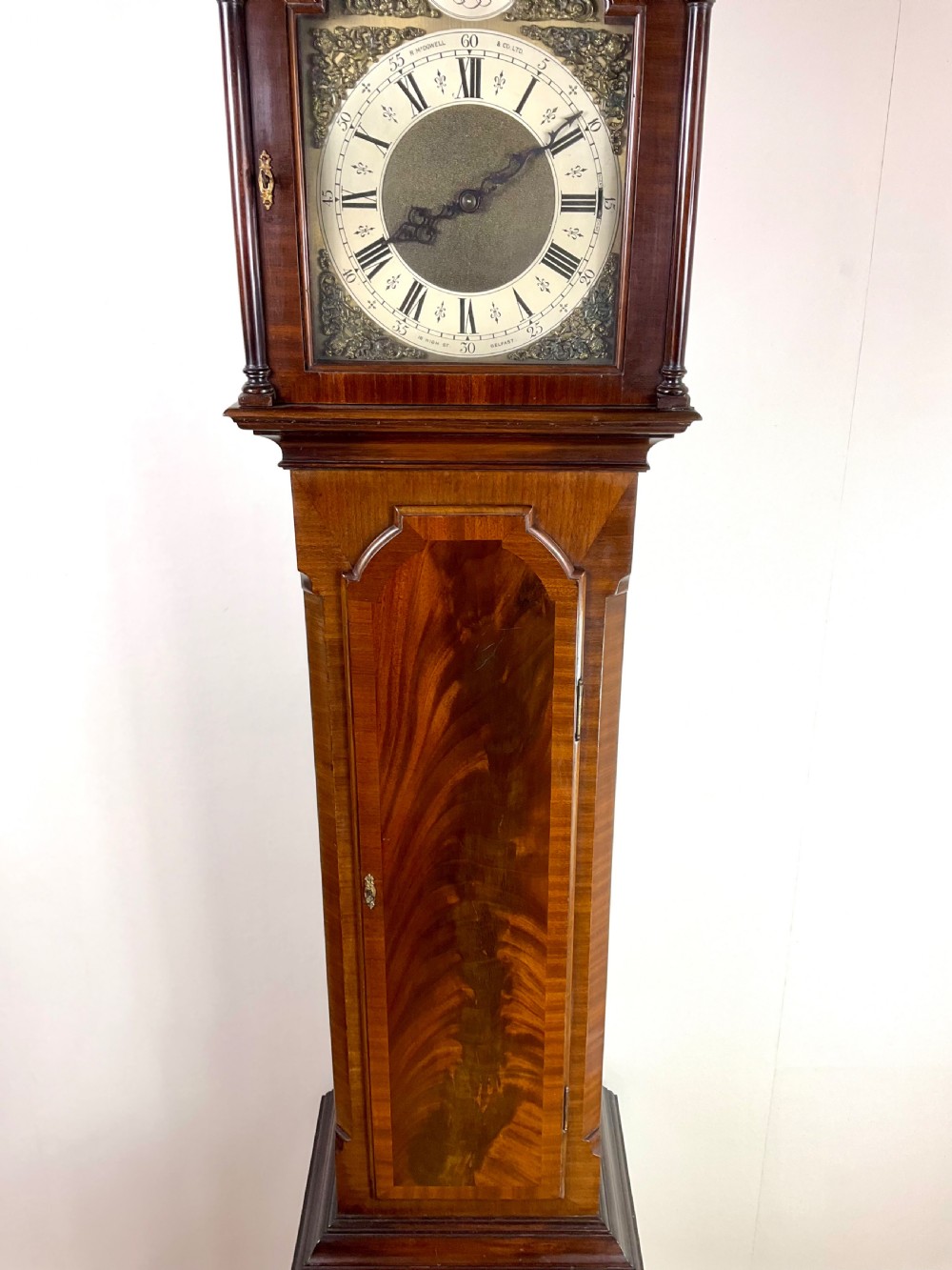 An antique mahogany longcase clock featuring a flame mahogany trunk door, arched hood, and brass dial with Roman numerals. View III