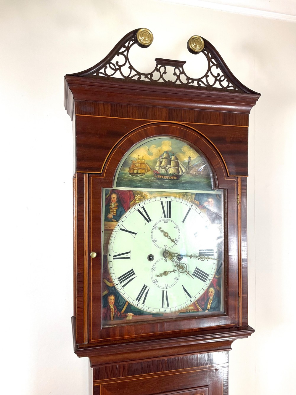 An upper hood section of a Georgian mahogany longcase clock featuring open fretwork pediments, brass finials, and a painted arch dial displaying a rocking ship automaton.