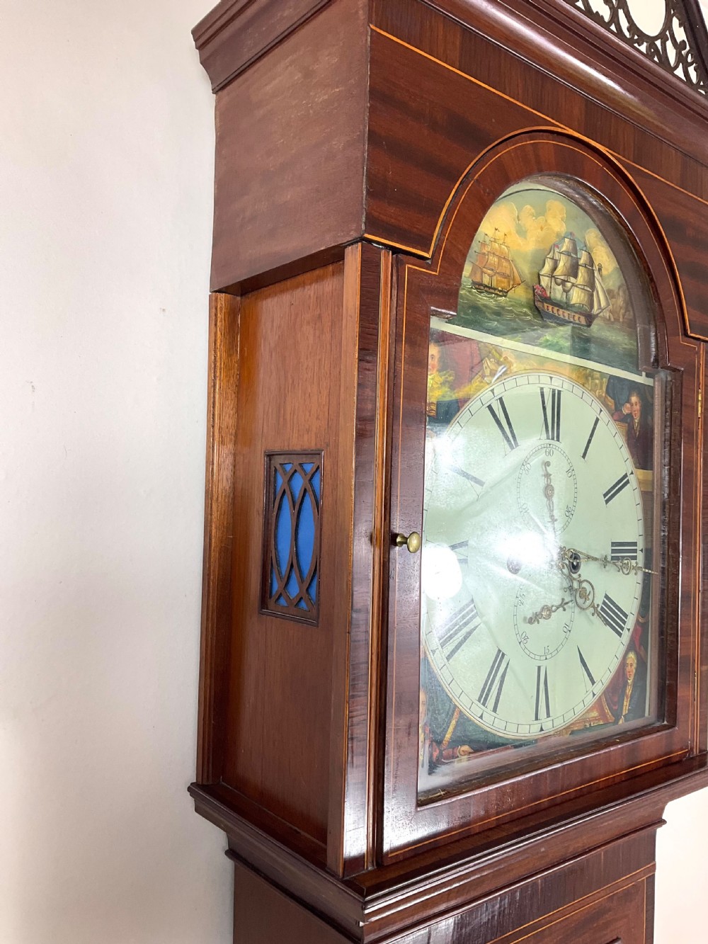 An upper hood section of a Georgian mahogany longcase clock featuring open fretwork pediments, brass finials, and a painted arch dial displaying a rocking ship automaton. View III