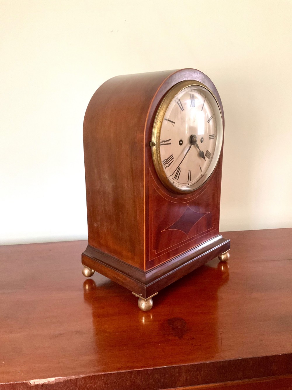 An English mahogany bracket clock featuring an arched top, white dial with Roman numerals, brass bezel, geometric marquetry inlay, and four brass ball feet.