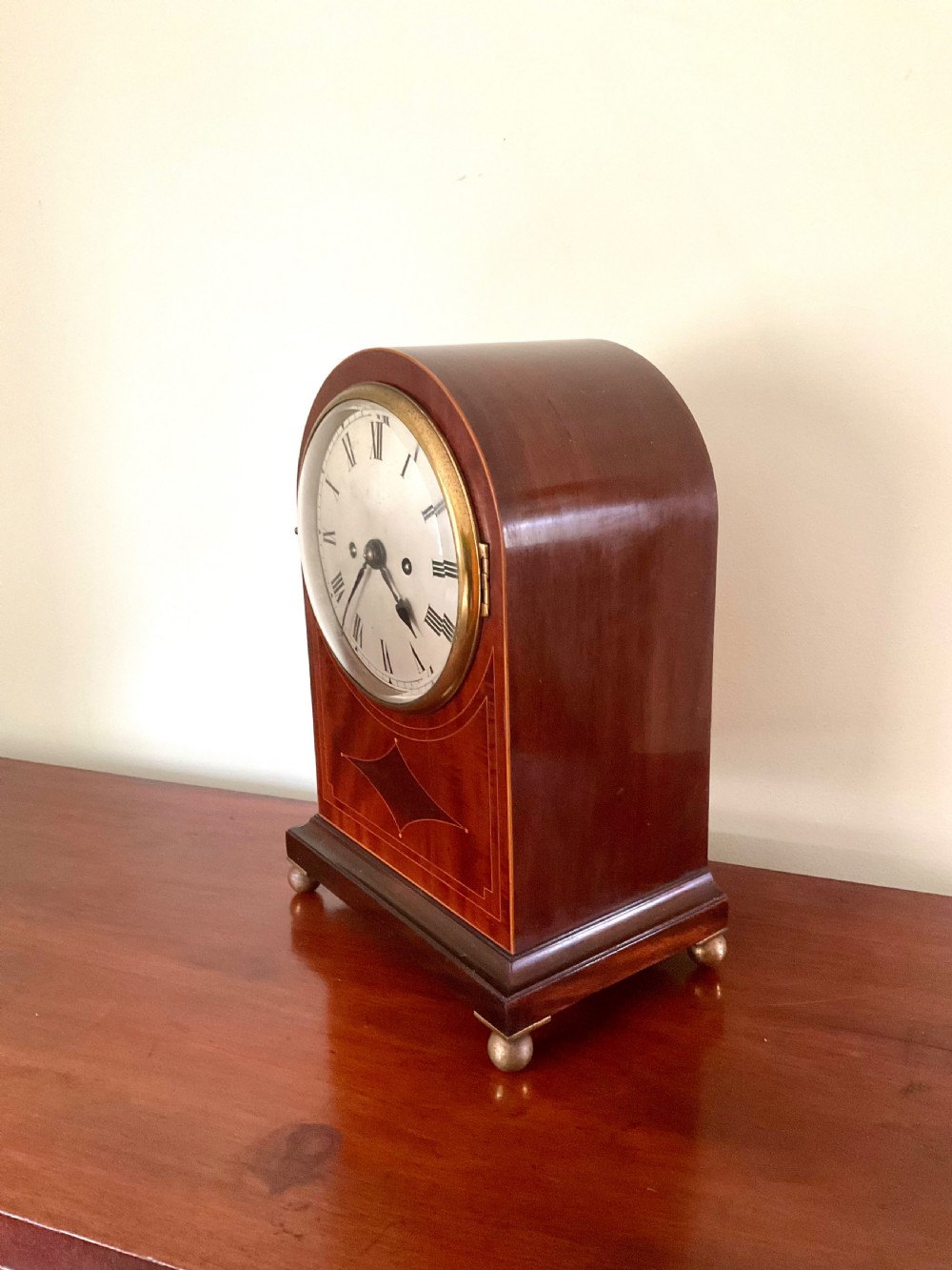 An English mahogany bracket clock featuring an arched top, white dial with Roman numerals, brass bezel, geometric marquetry inlay, and four brass ball feet. View II