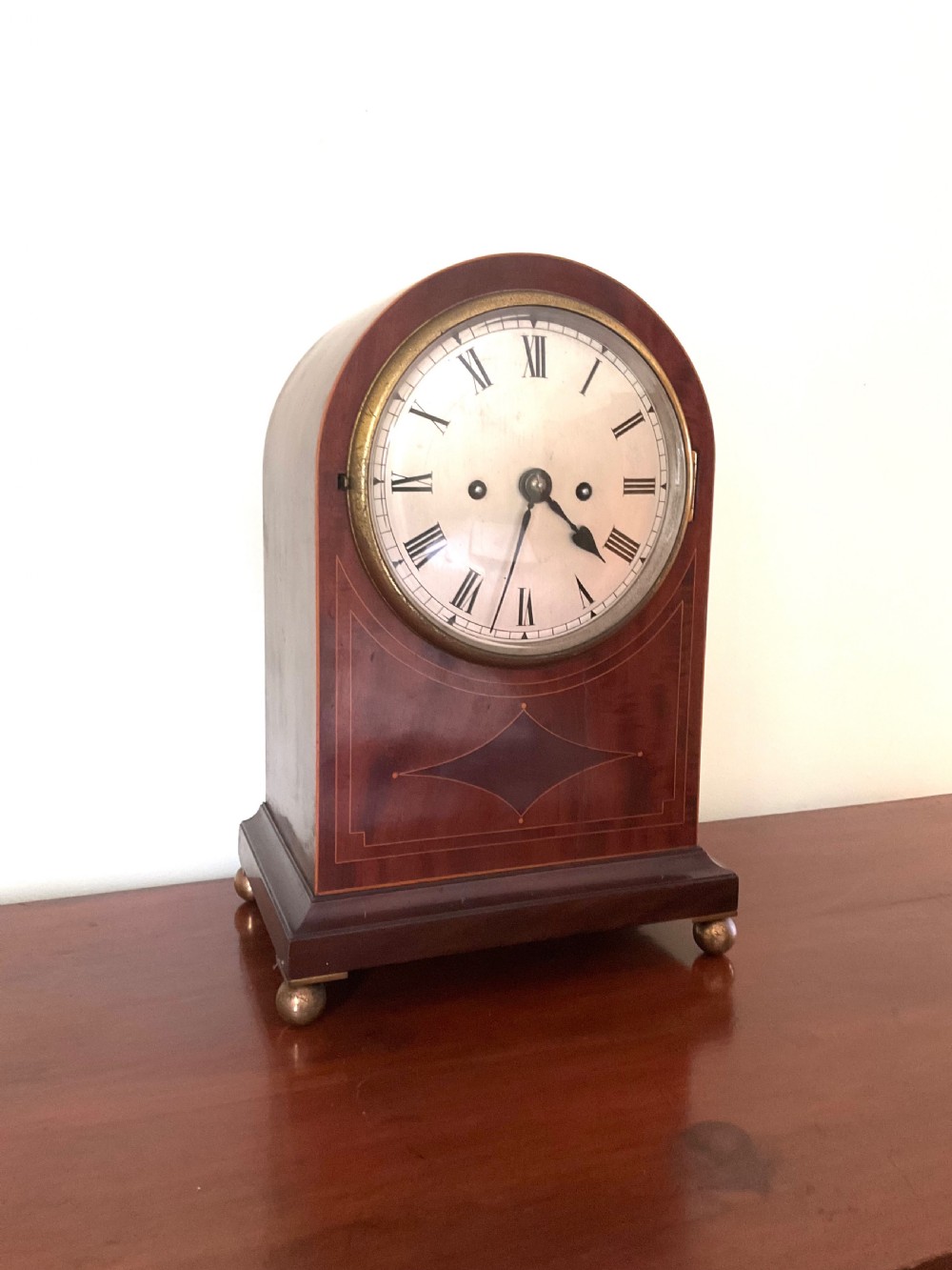 An English mahogany bracket clock featuring an arched top, white dial with Roman numerals, brass bezel, geometric marquetry inlay, and four brass ball feet. View III