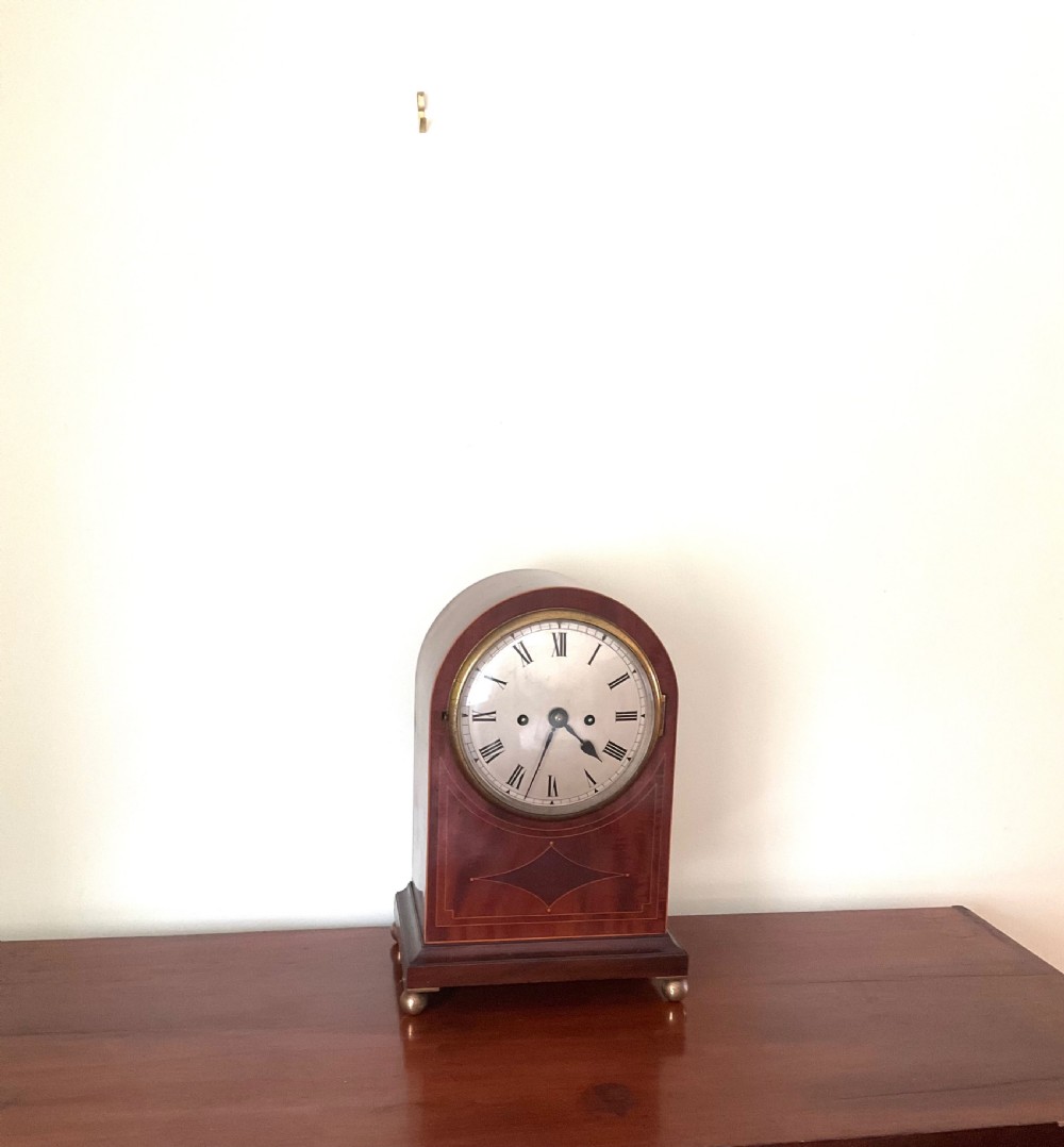 An English mahogany bracket clock featuring an arched top, white dial with Roman numerals, brass bezel, geometric marquetry inlay, and four brass ball feet. View IV