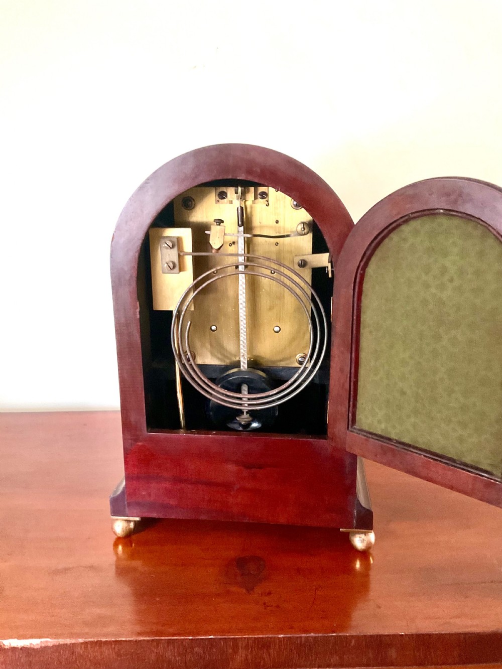 An English mahogany bracket clock featuring an arched top, white dial with Roman numerals, brass bezel, geometric marquetry inlay, and four brass ball feet. View VI