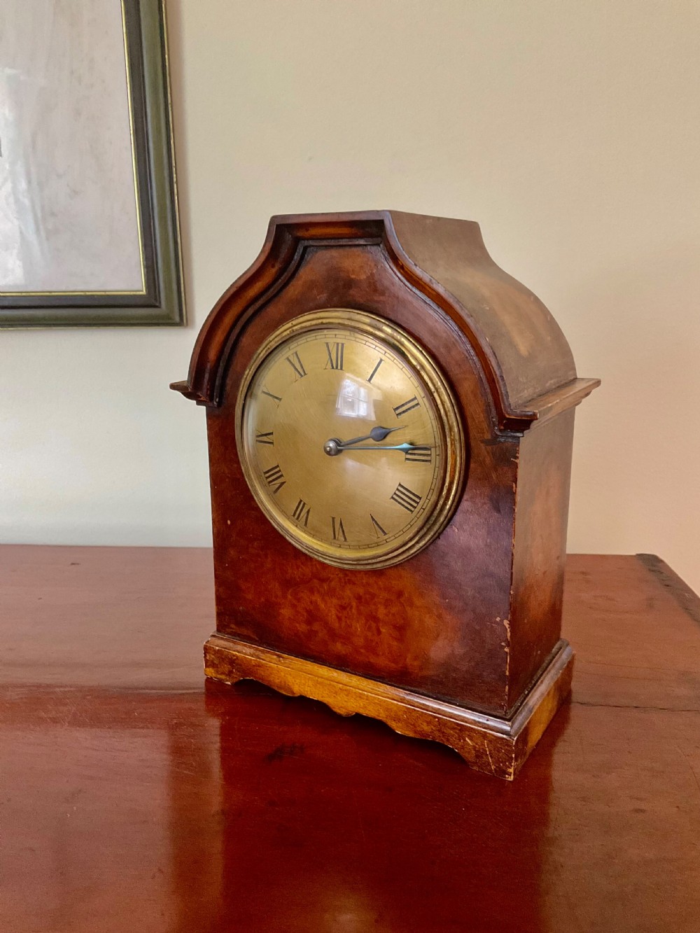 A small figured walnut mantel clock featuring an arched timber case, a brass-framed dial, and black Roman numerals.