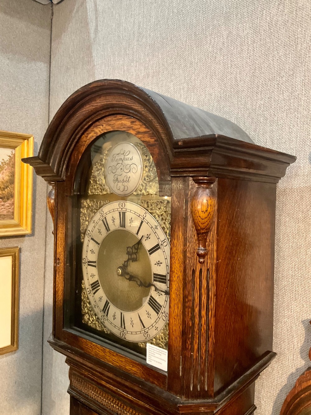 A tall dark oak grandfather clock featuring an arched hood, brass dial with spandrels, and a carved oval motif on the central door. View III