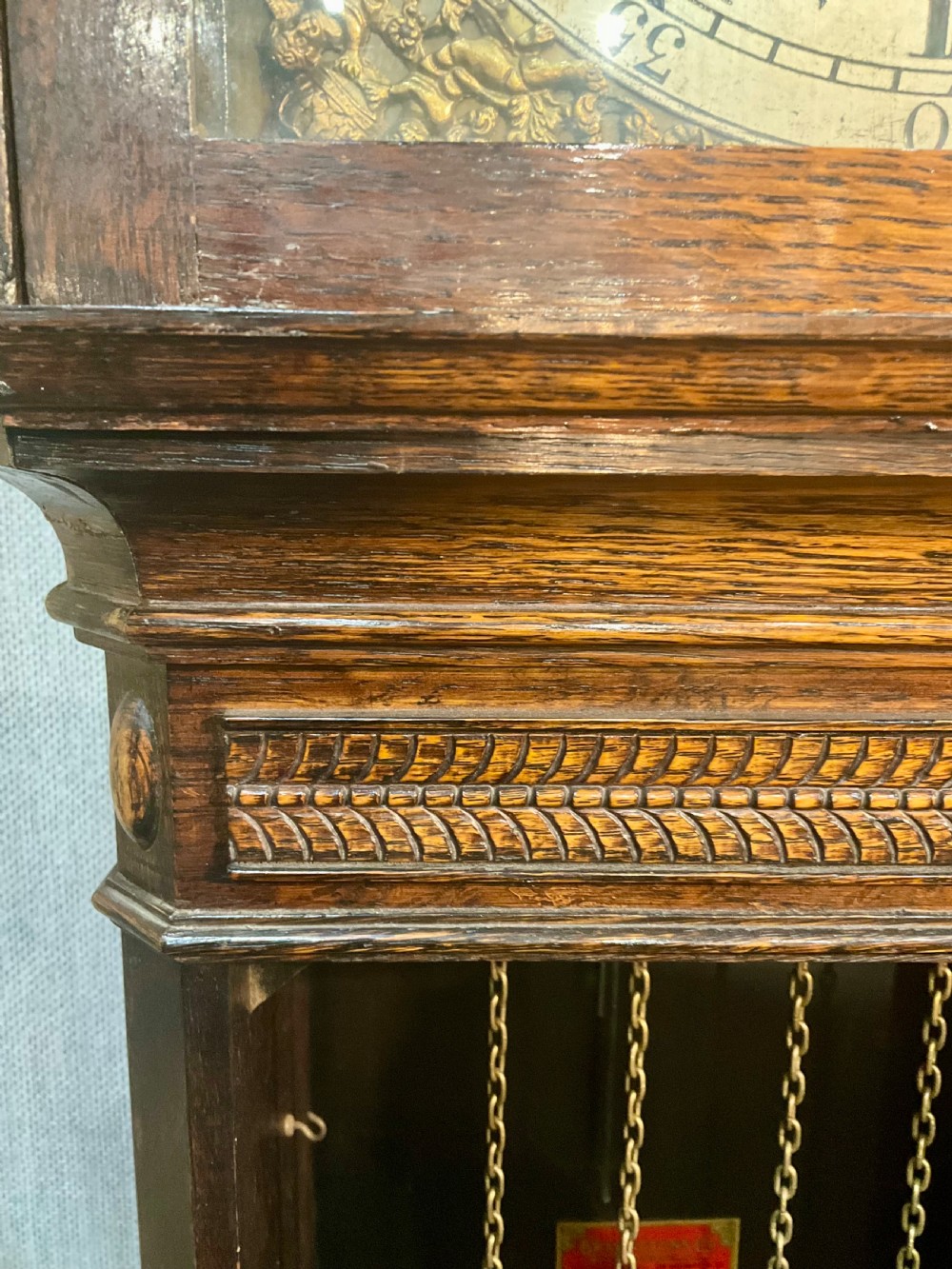 A tall dark oak grandfather clock featuring an arched hood, brass dial with spandrels, and a carved oval motif on the central door. View IV