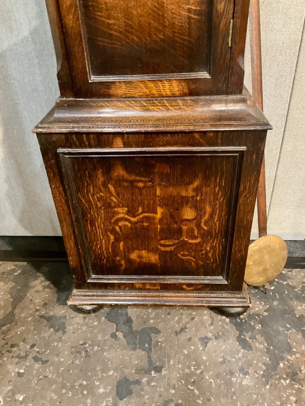 A tall dark oak grandfather clock featuring an arched hood, brass dial with spandrels, and a carved oval motif on the central door. View V
