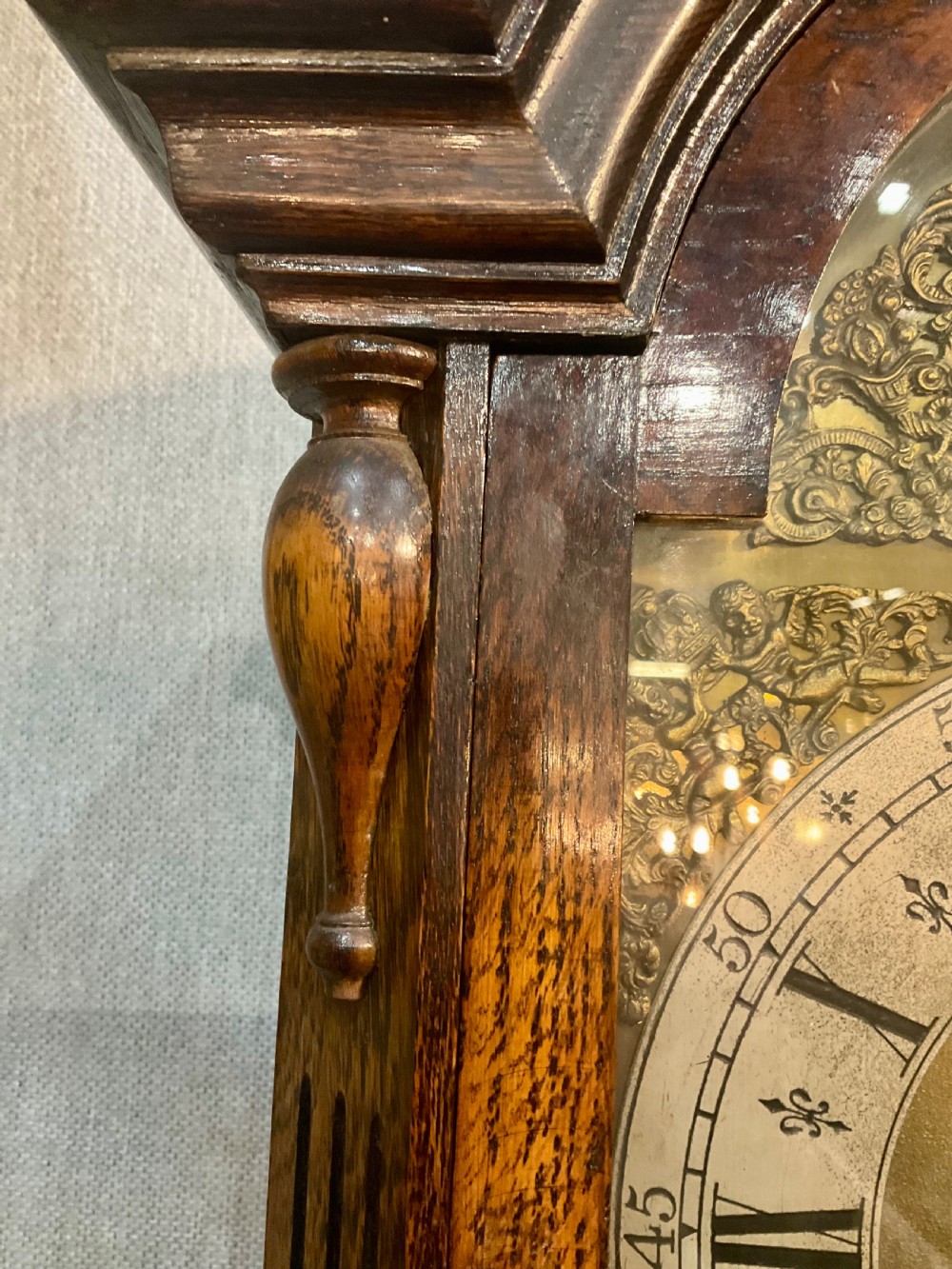 A tall dark oak grandfather clock featuring an arched hood, brass dial with spandrels, and a carved oval motif on the central door. View VII