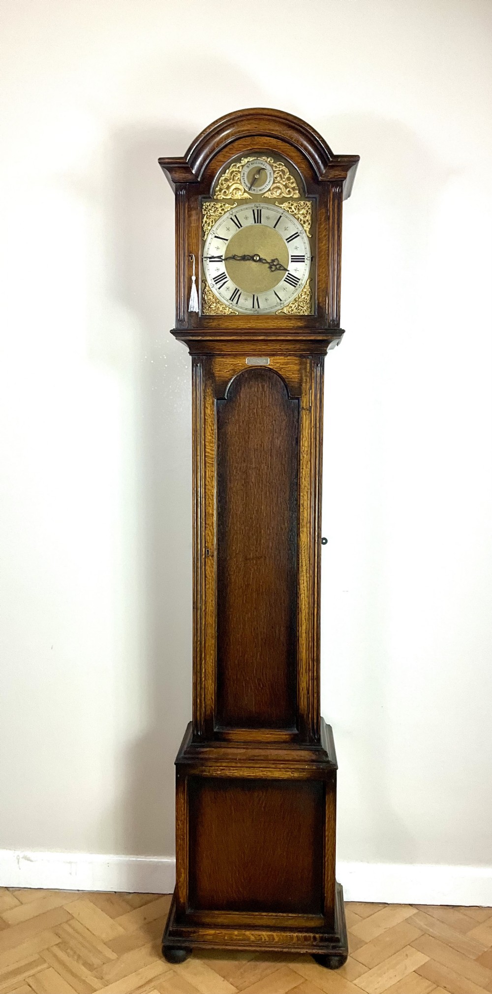 An antique dark oak longcase grandfather clock featuring an arched hood, brass dial with silvered chapter ring, and a tall panelled trunk.