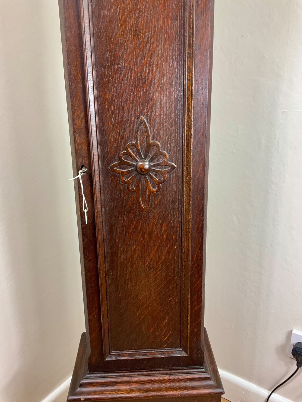 A small dark oak grandmother longcase clock featuring an arched hood, a brass dial with Roman numerals, and a carved floral motif on the trunk door. View III