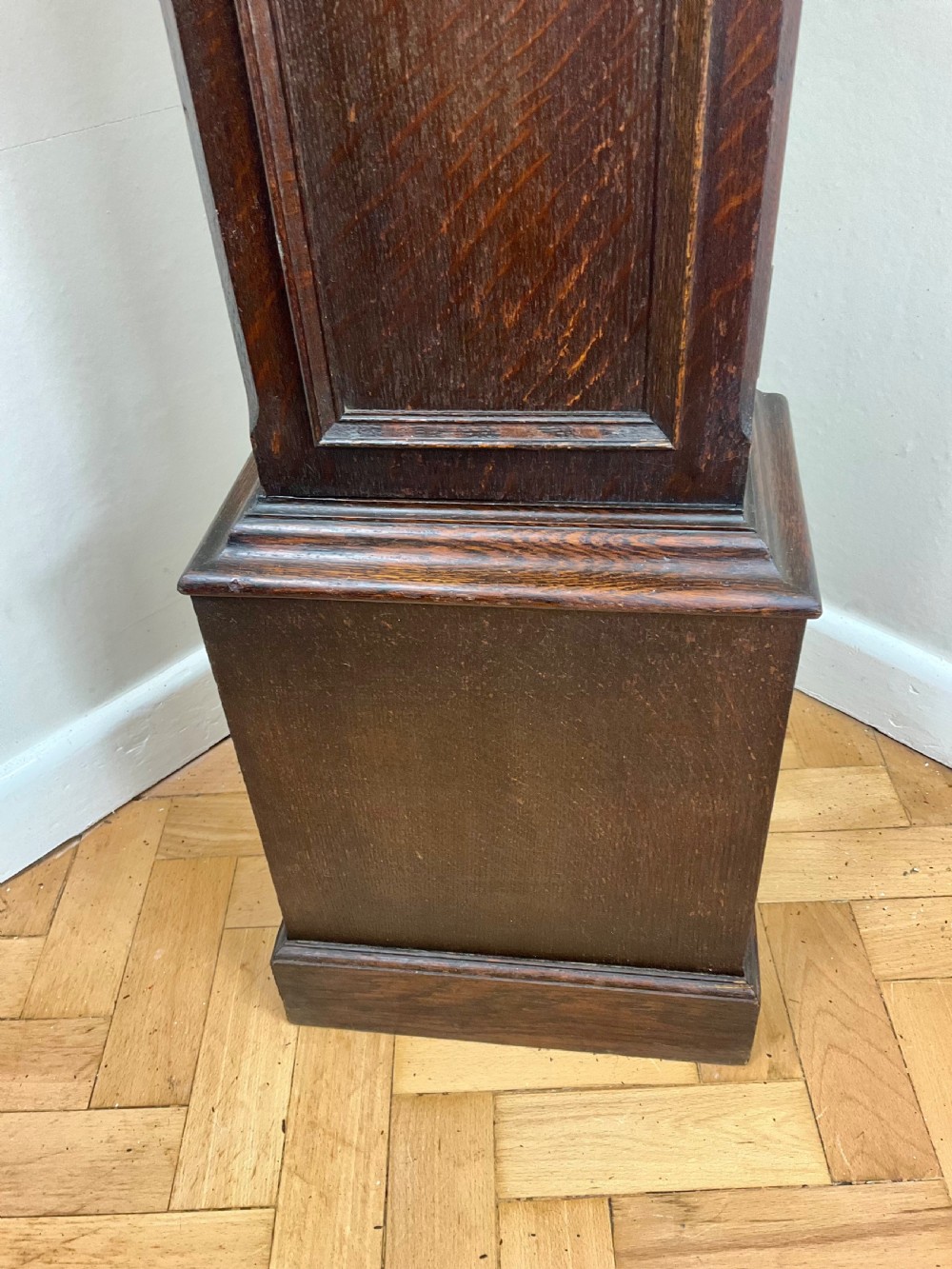 A small dark oak grandmother longcase clock featuring an arched hood, a brass dial with Roman numerals, and a carved floral motif on the trunk door. View IV