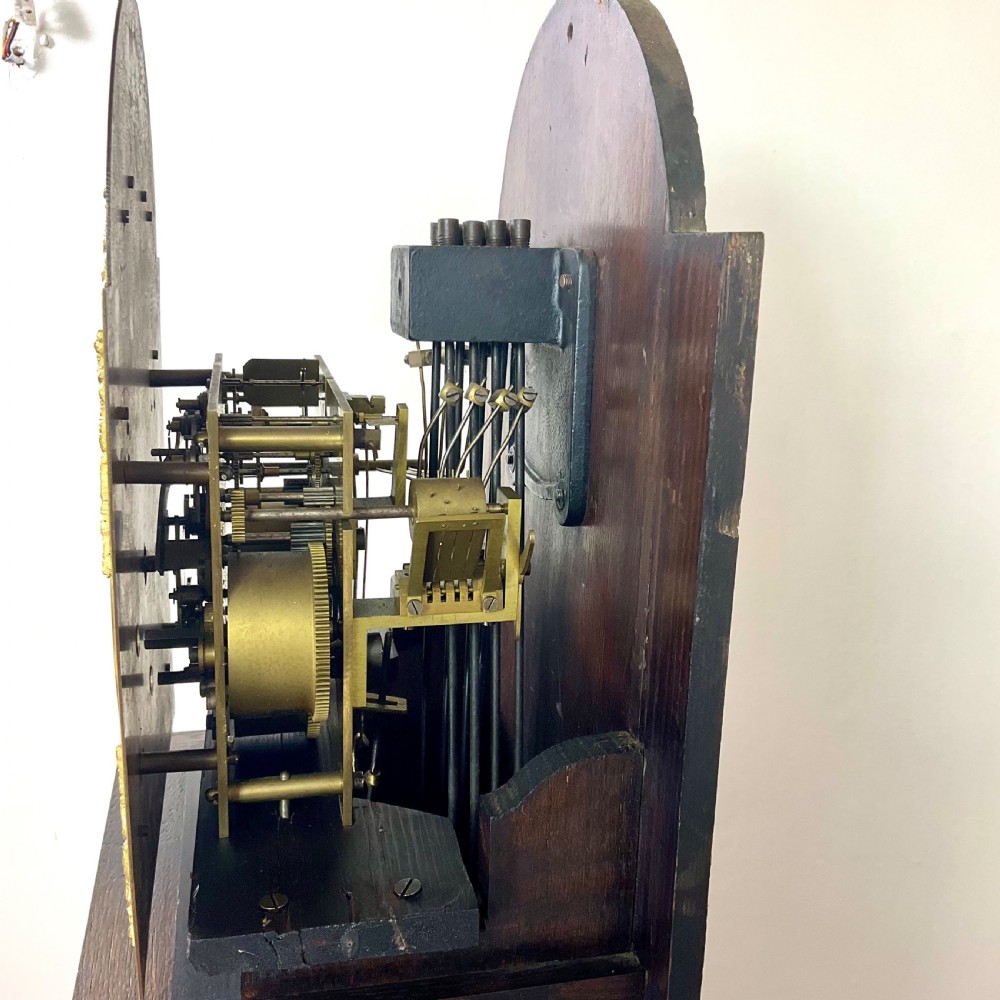 A small dark oak grandmother longcase clock featuring an arched hood, a brass dial with Roman numerals, and a carved floral motif on the trunk door. View VIII