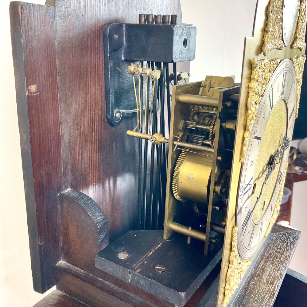 A small dark oak grandmother longcase clock featuring an arched hood, a brass dial with Roman numerals, and a carved floral motif on the trunk door. View IX