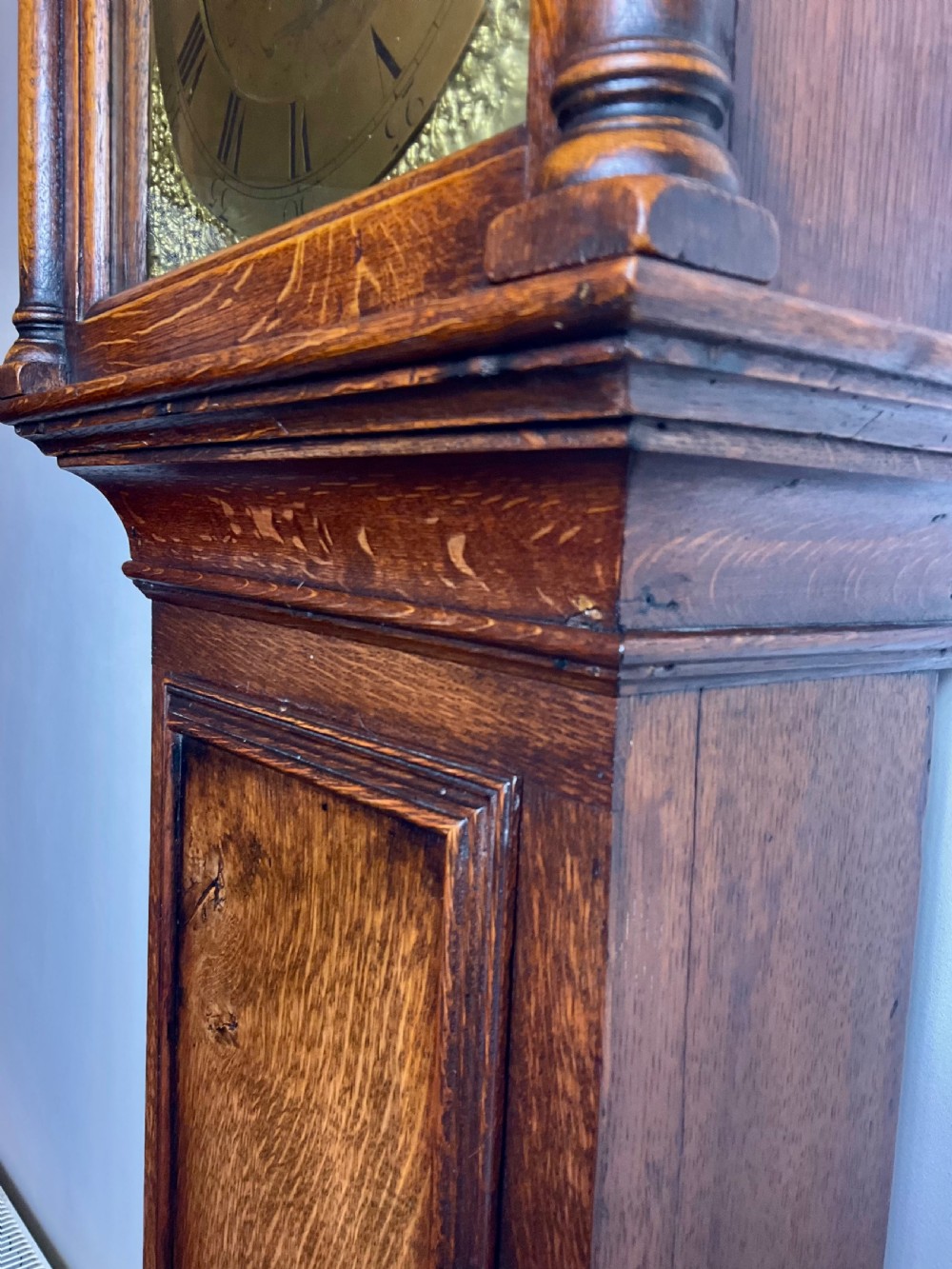 An Early Georgian oak longcase clock featuring a square brass dial with spandrels, flanked by turned columns, and a tall wooden trunk set on bracket feet. View II