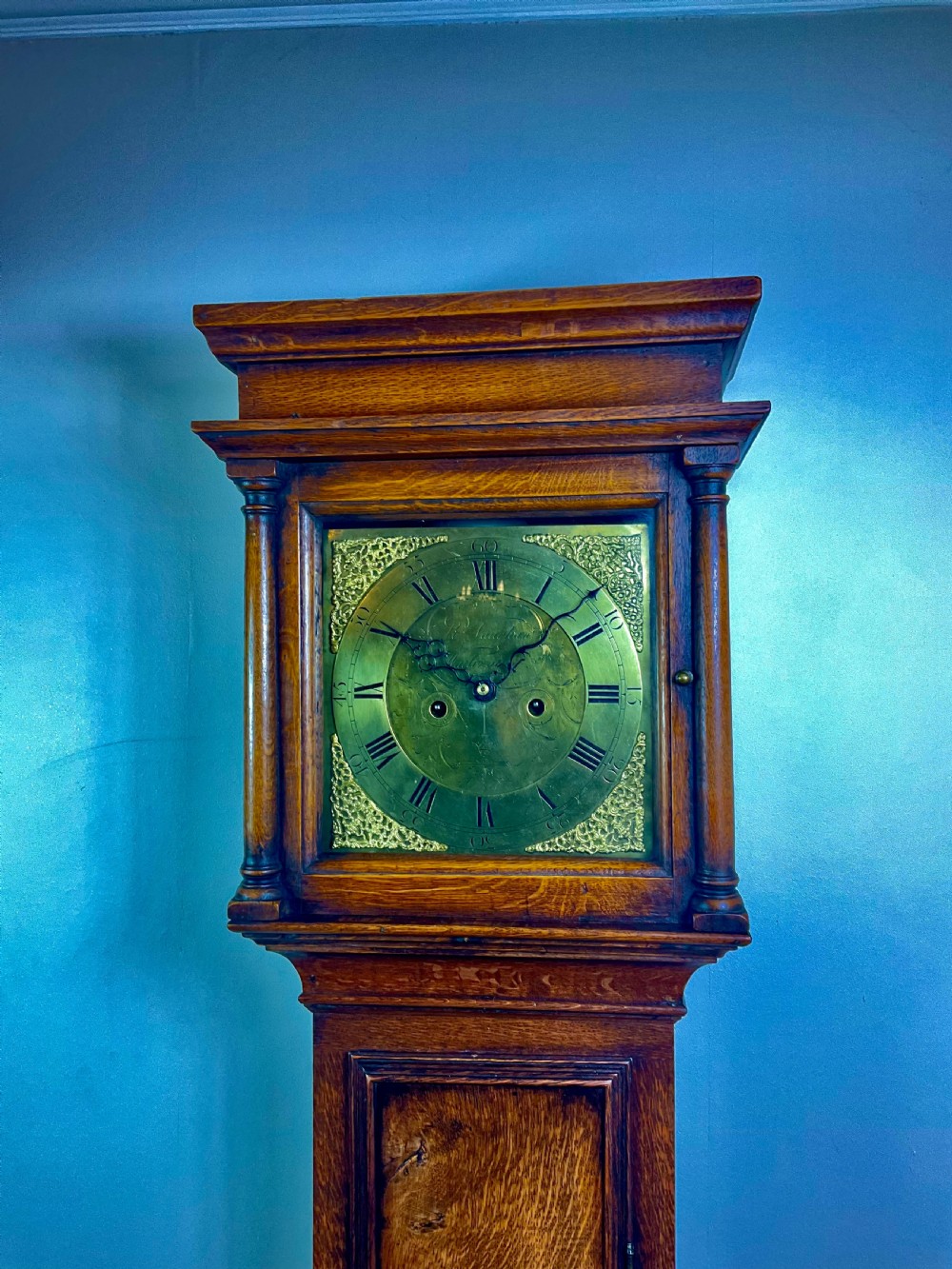 An Early Georgian oak longcase clock featuring a square brass dial with spandrels, flanked by turned columns, and a tall wooden trunk set on bracket feet. View V