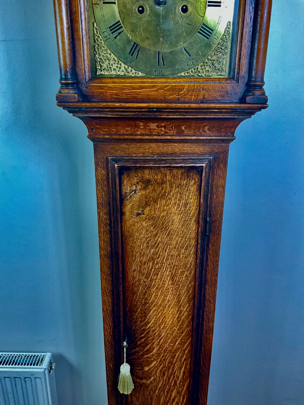An Early Georgian oak longcase clock featuring a square brass dial with spandrels, flanked by turned columns, and a tall wooden trunk set on bracket feet. View XIII