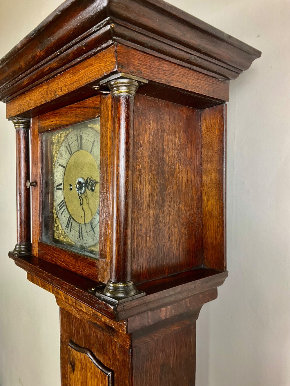 A slim Georgian oak longcase clock featuring a square brass dial with spandrels, fluted hood columns, a hinged trunk door, and a plinth base. View V