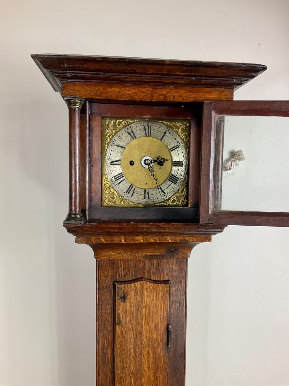 A slim Georgian oak longcase clock featuring a square brass dial with spandrels, fluted hood columns, a hinged trunk door, and a plinth base. View IX