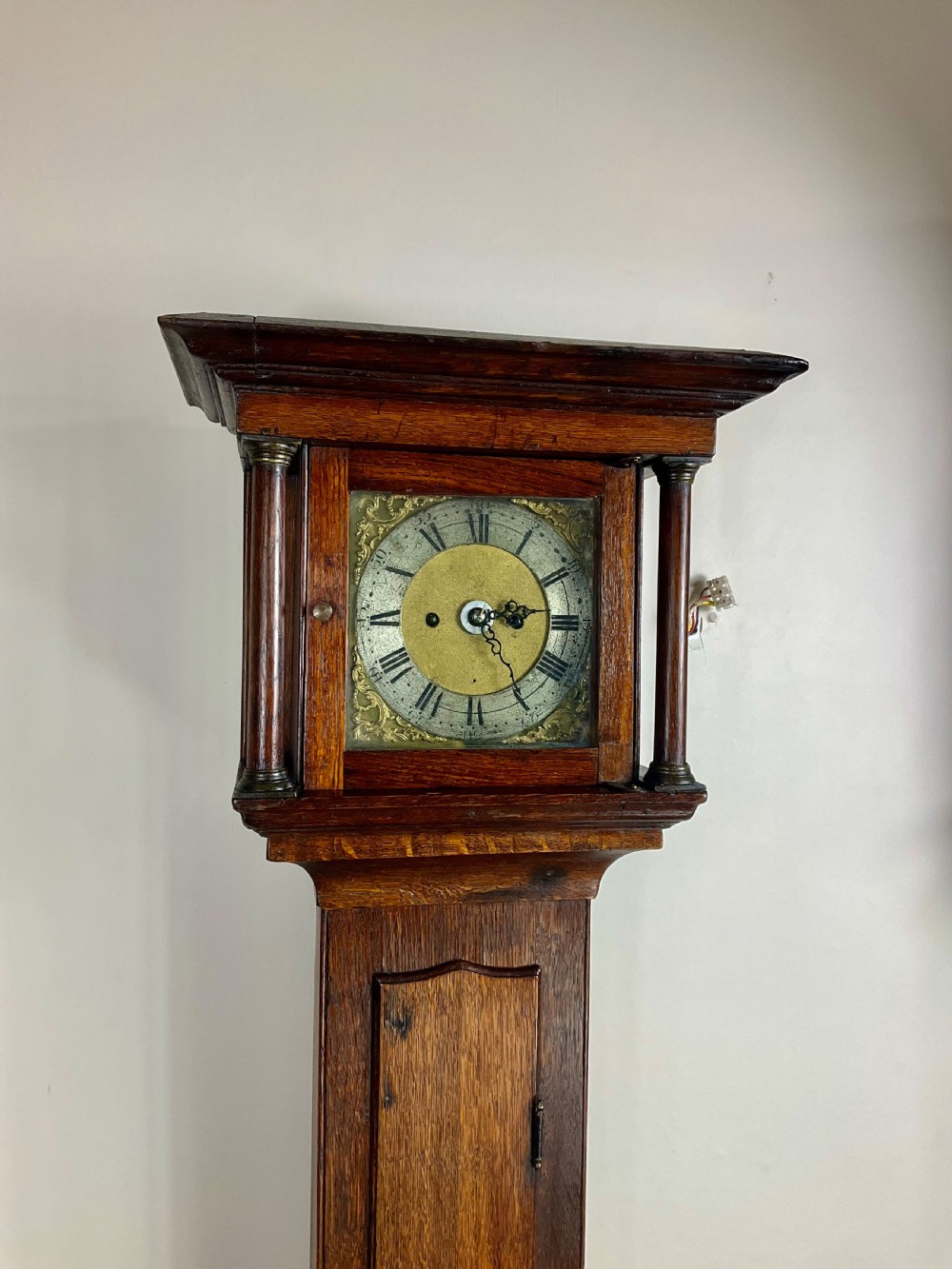 A slim Georgian oak longcase clock featuring a square brass dial with spandrels, fluted hood columns, a hinged trunk door, and a plinth base. View X