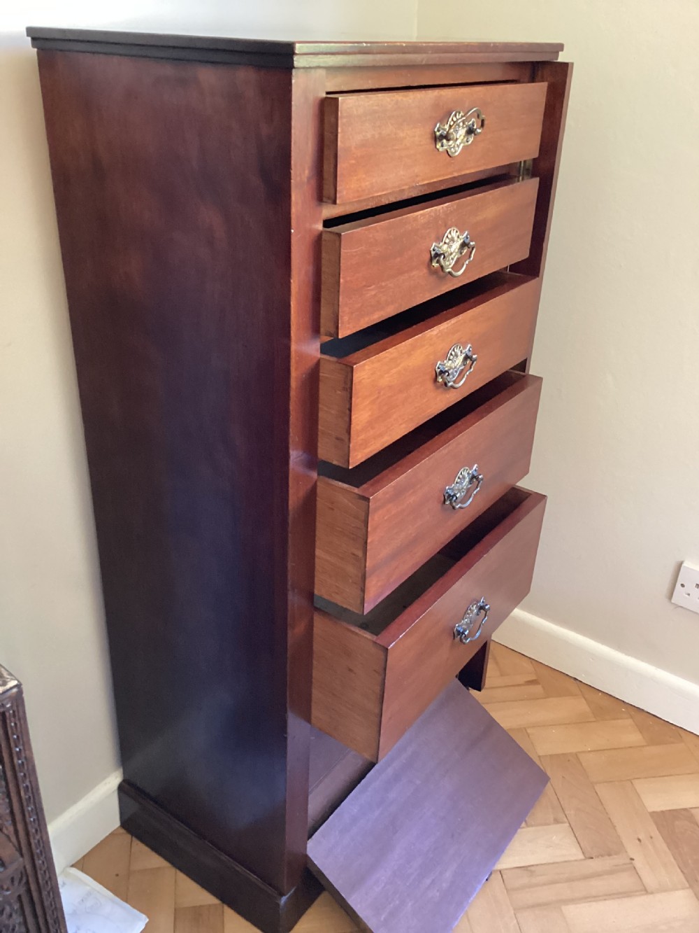 A tall, narrow mahogany Wellington chest featuring six drawers with ornate brass handles and a hinged locking side bar on a plinth base. View III