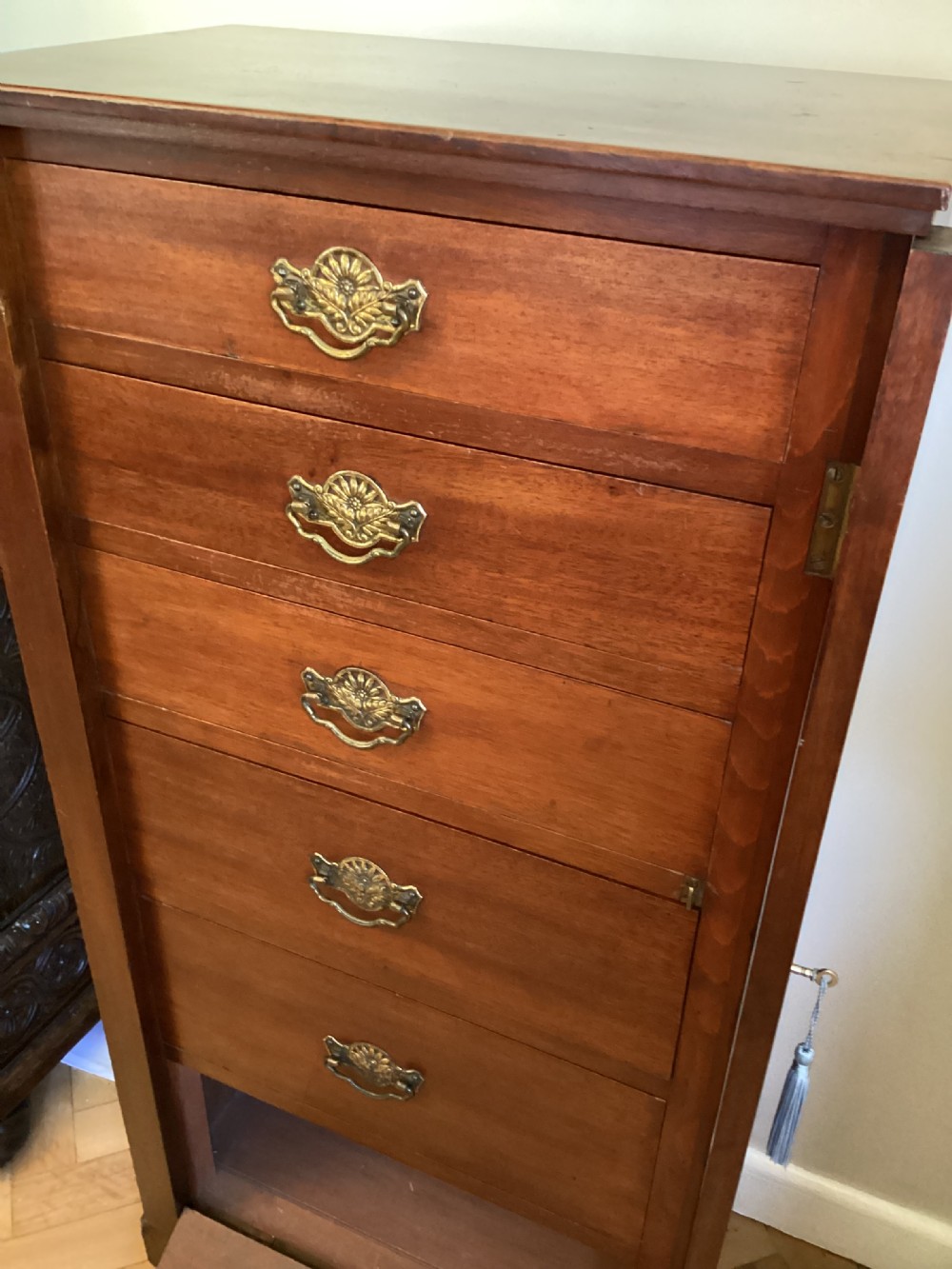 A tall, narrow mahogany Wellington chest featuring six drawers with ornate brass handles and a hinged locking side bar on a plinth base. View V