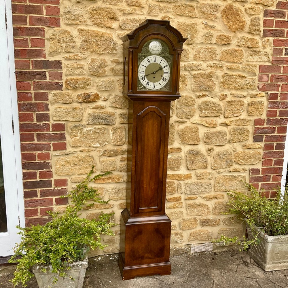 A floor-standing walnut grandmother longcase clock featuring an arched hood, brass dial with Roman numerals, long trunk door, and stepped plinth base.