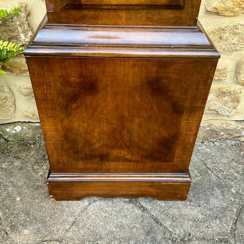 A floor-standing walnut grandmother longcase clock featuring an arched hood, brass dial with Roman numerals, long trunk door, and stepped plinth base. View IV