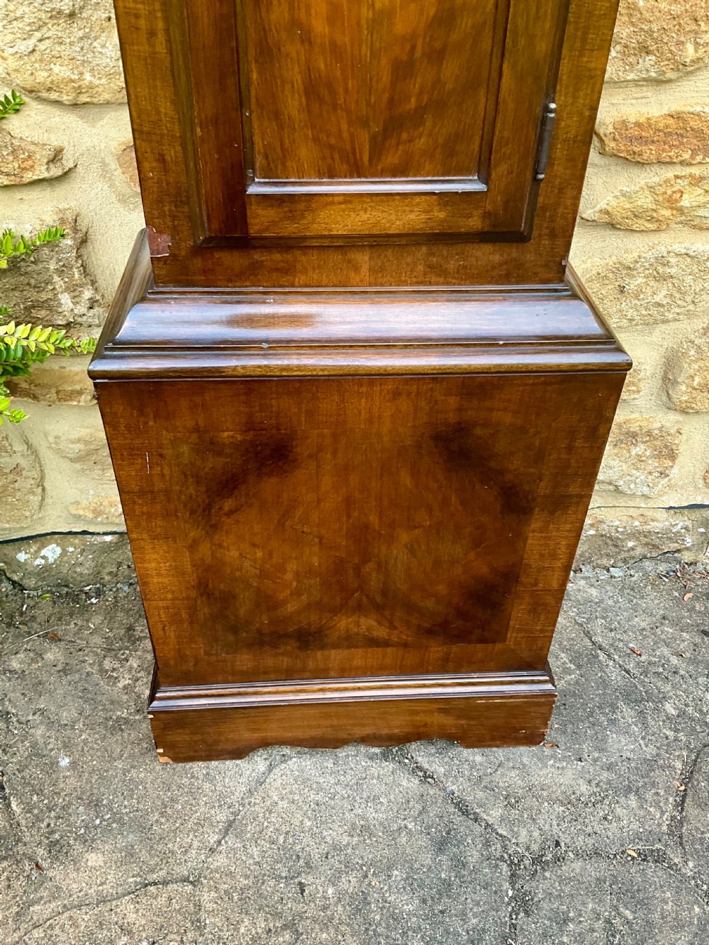 A floor-standing walnut grandmother longcase clock featuring an arched hood, brass dial with Roman numerals, long trunk door, and stepped plinth base. View V