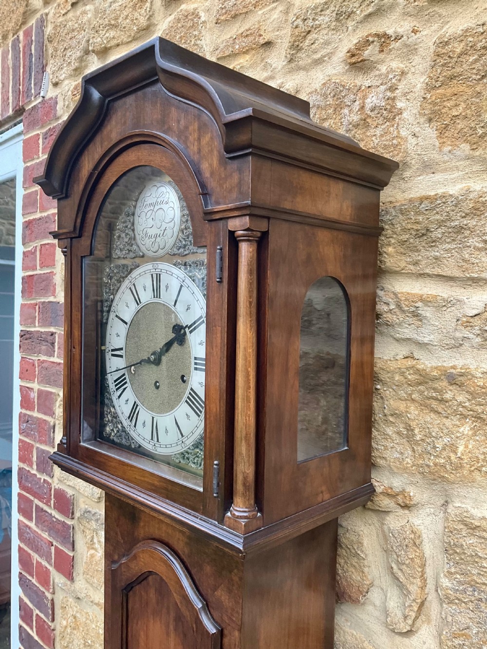 A floor-standing walnut grandmother longcase clock featuring an arched hood, brass dial with Roman numerals, long trunk door, and stepped plinth base. View VII