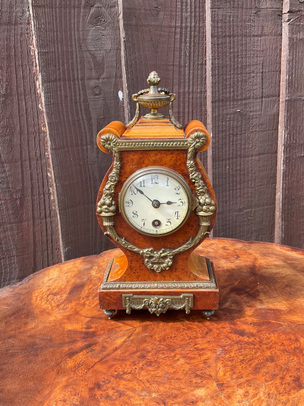 A small lyre-shaped mantel clock featuring figured burr walnut veneer, gilt ormolu swags, an urn finial, and an enamelled dial with Arabic numerals.