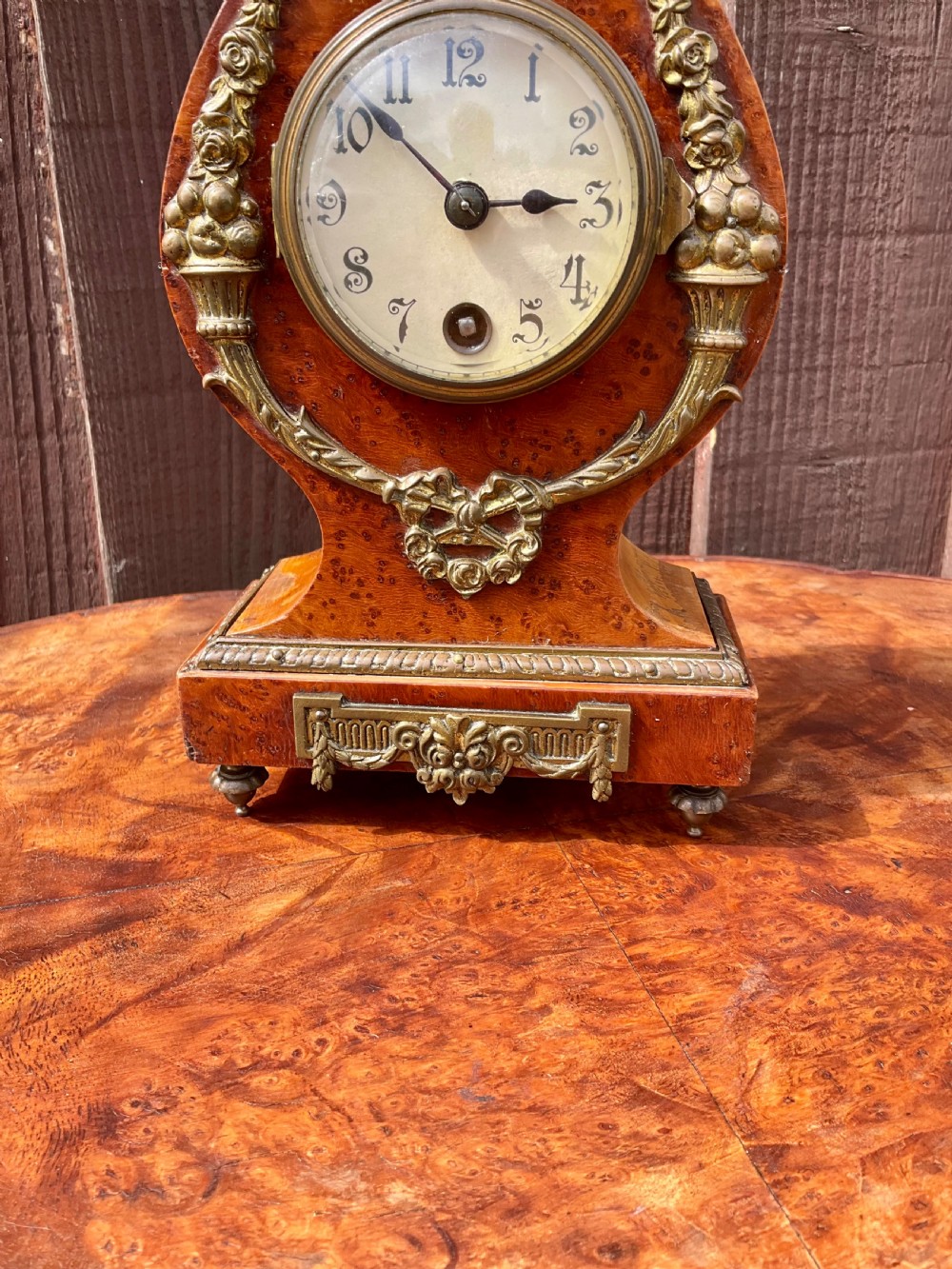 A small lyre-shaped mantel clock featuring figured burr walnut veneer, gilt ormolu swags, an urn finial, and an enamelled dial with Arabic numerals. View VI