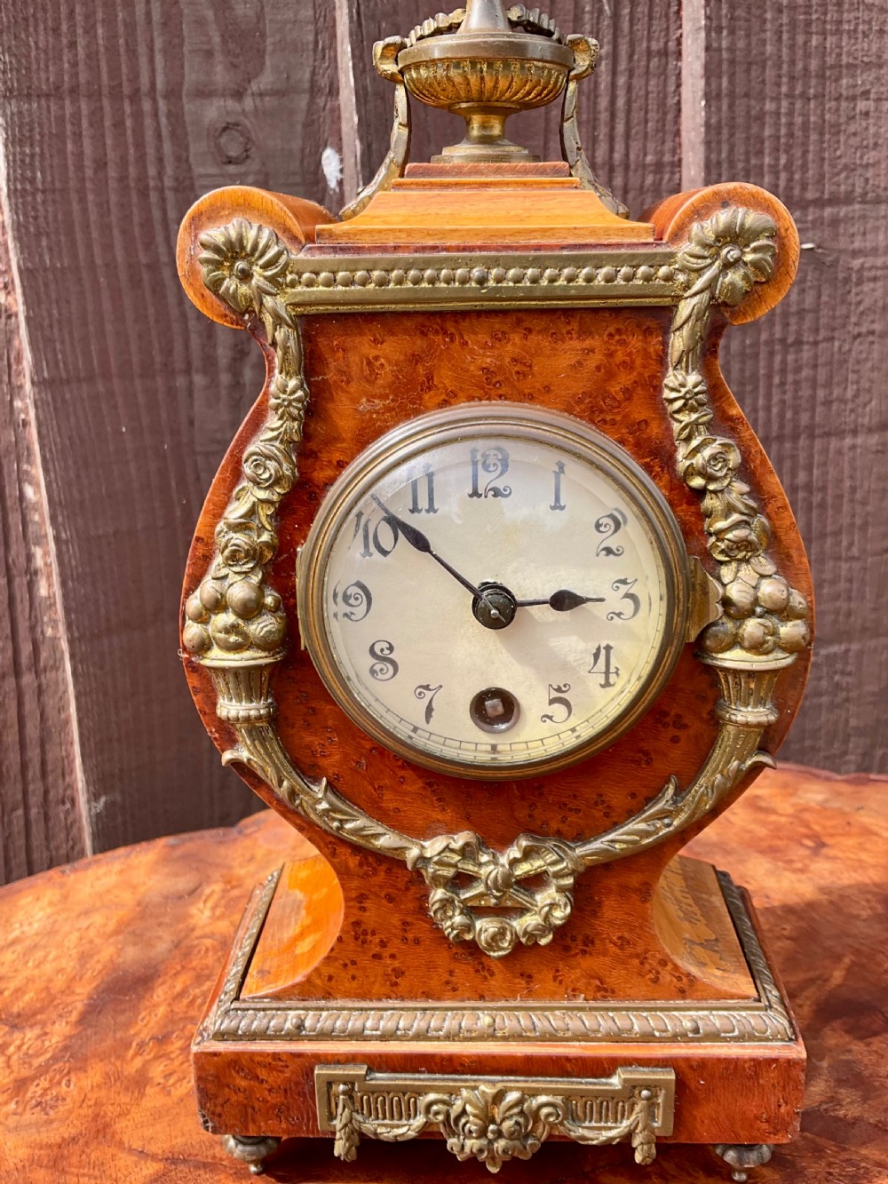 A small lyre-shaped mantel clock featuring figured burr walnut veneer, gilt ormolu swags, an urn finial, and an enamelled dial with Arabic numerals. View IX