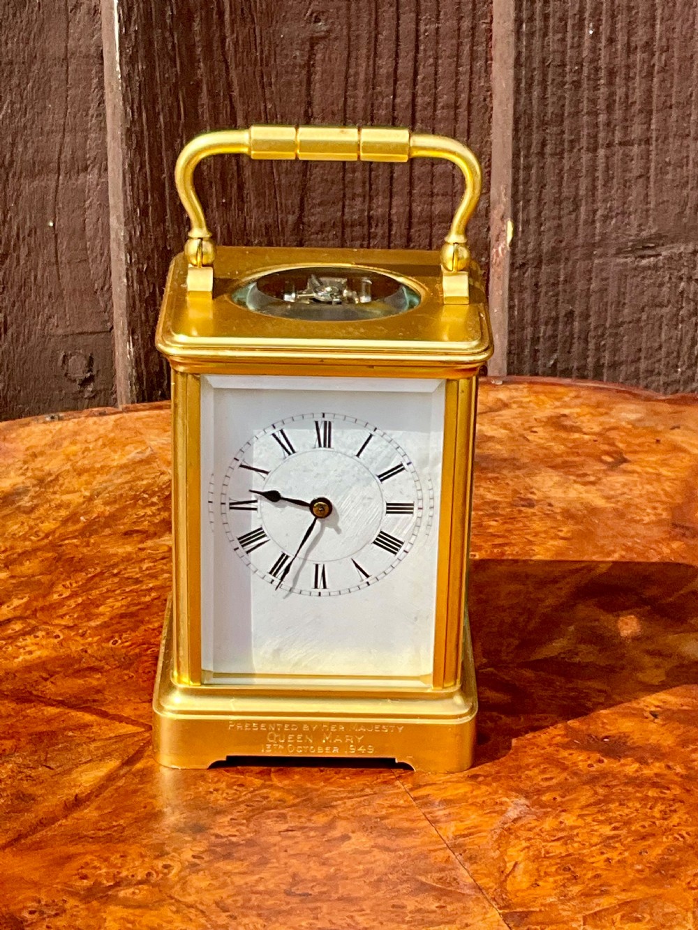A rectangular gilt brass carriage clock featuring a top carrying handle, a circular top window displaying the balance wheel, and a white dial with Roman numerals.