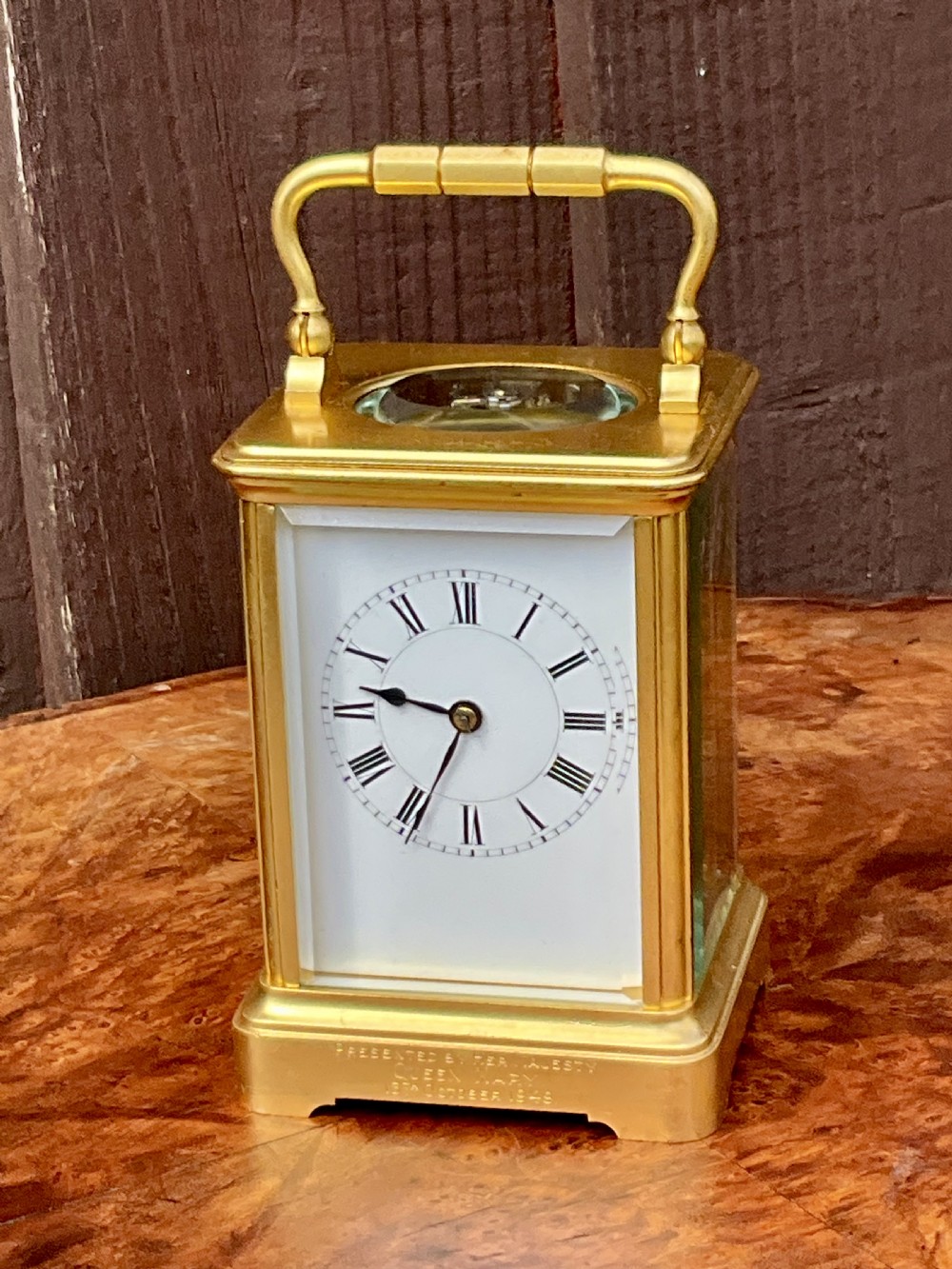 A rectangular gilt brass carriage clock featuring a top carrying handle, a circular top window displaying the balance wheel, and a white dial with Roman numerals. View IV