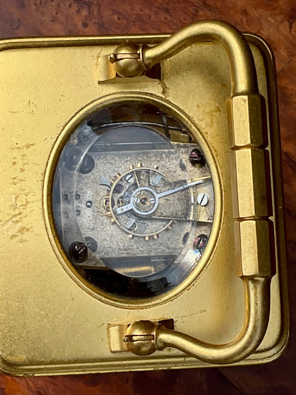 A rectangular gilt brass carriage clock featuring a top carrying handle, a circular top window displaying the balance wheel, and a white dial with Roman numerals. View V