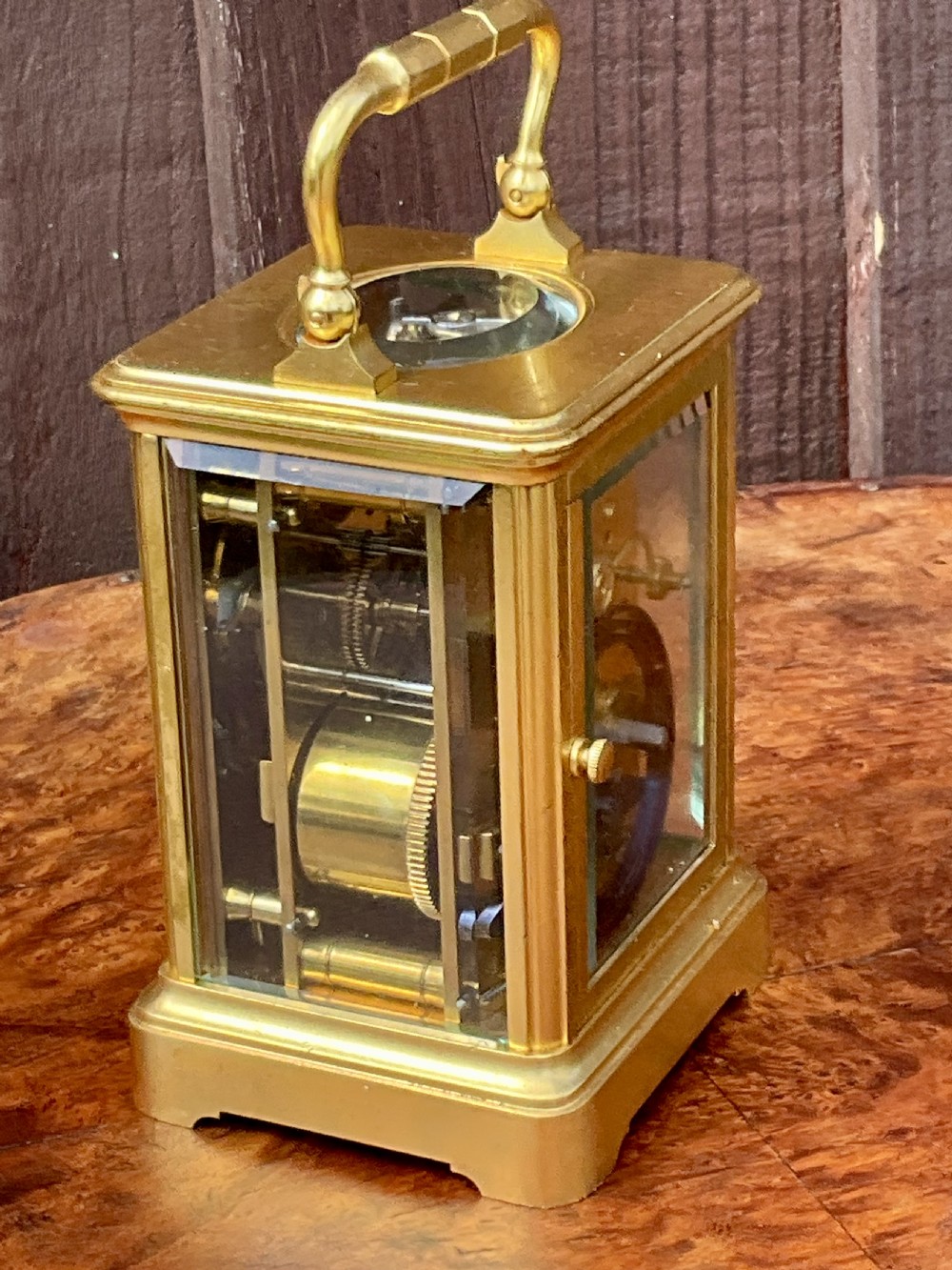 A rectangular gilt brass carriage clock featuring a top carrying handle, a circular top window displaying the balance wheel, and a white dial with Roman numerals. View VI