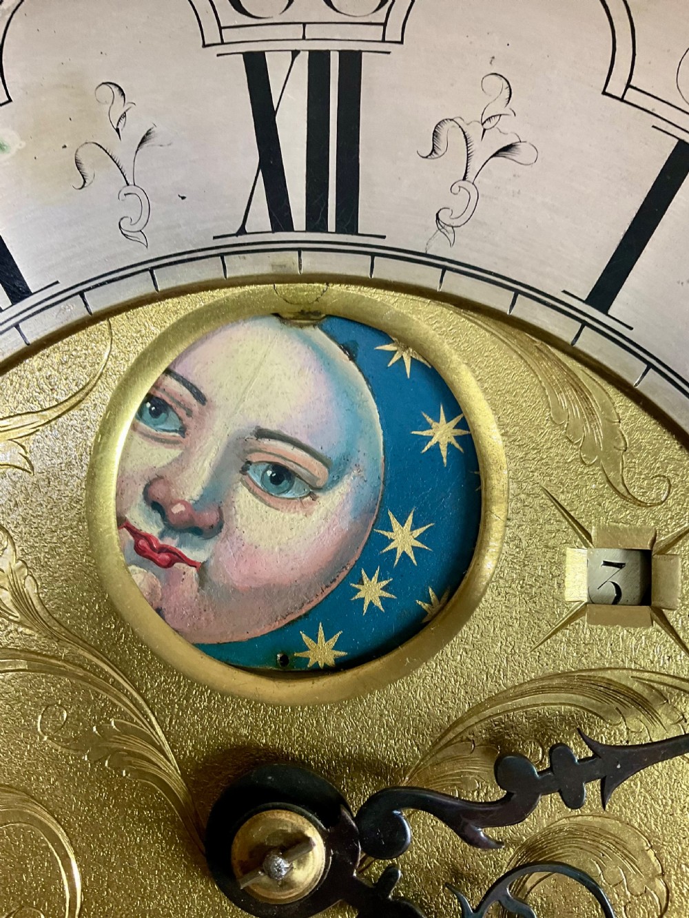 A close-up detail of an antique brass clock dial featuring a hand-painted penny moon face set against a blue sky with gold stars.