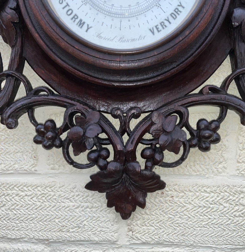 A 19th-century Black Forest aneroid barometer featuring a carved dark wood frame with leaf and acorn motifs surrounding a circular white ceramic dial. View VI